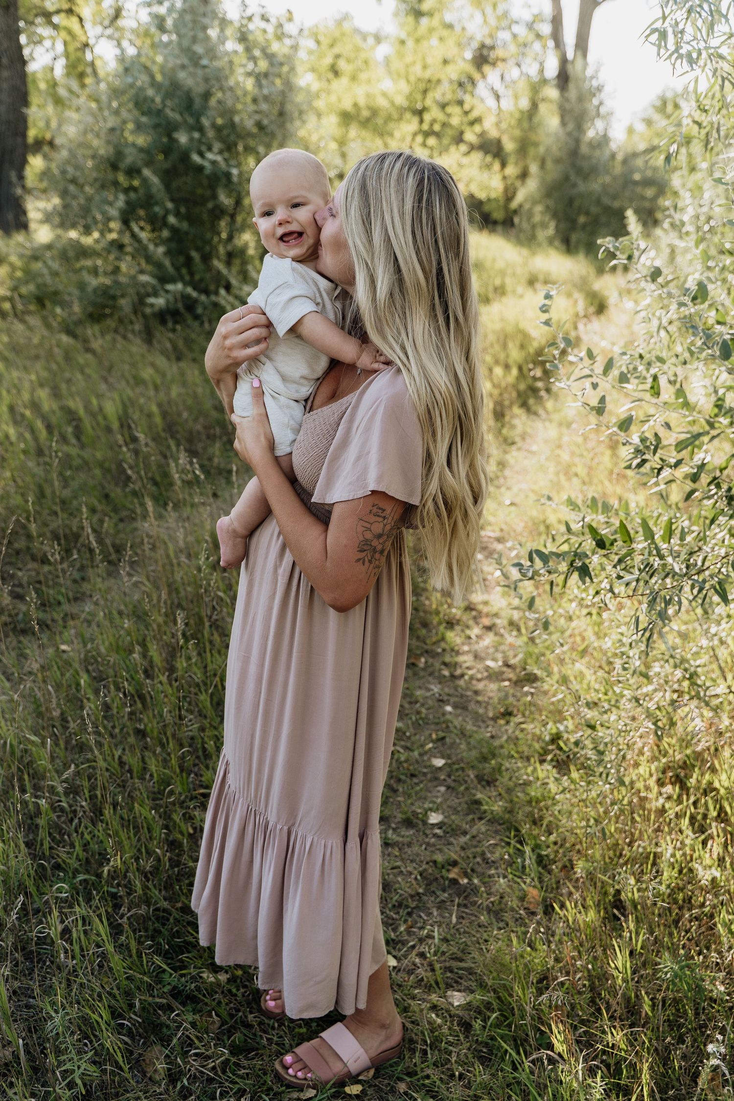 Woman in maxi dress holding smiling baby outdoors on a lush green nature path.