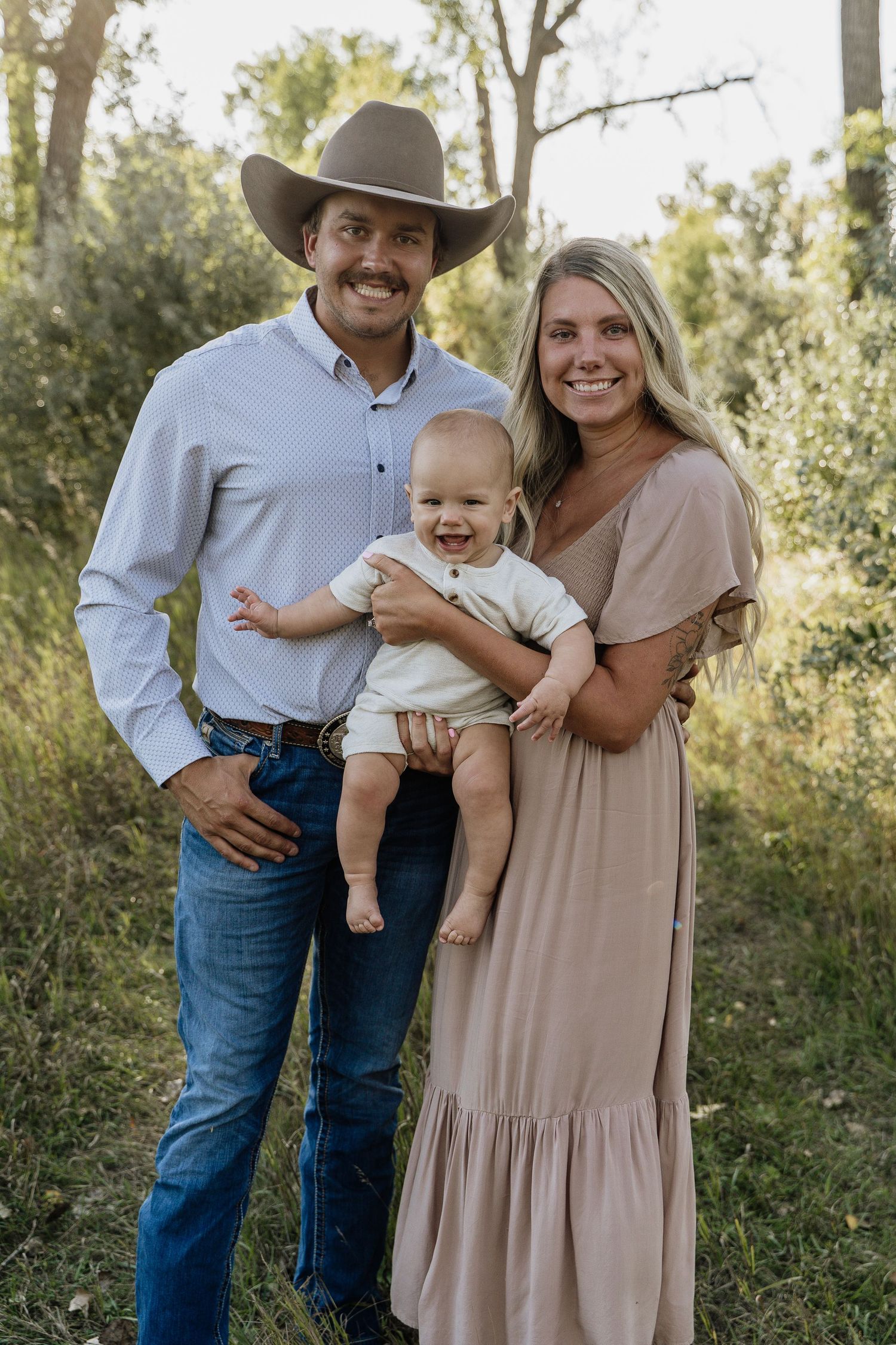 Happy young family with baby in outdoor nature portrait, man in cowboy hat and woman in beige dress.