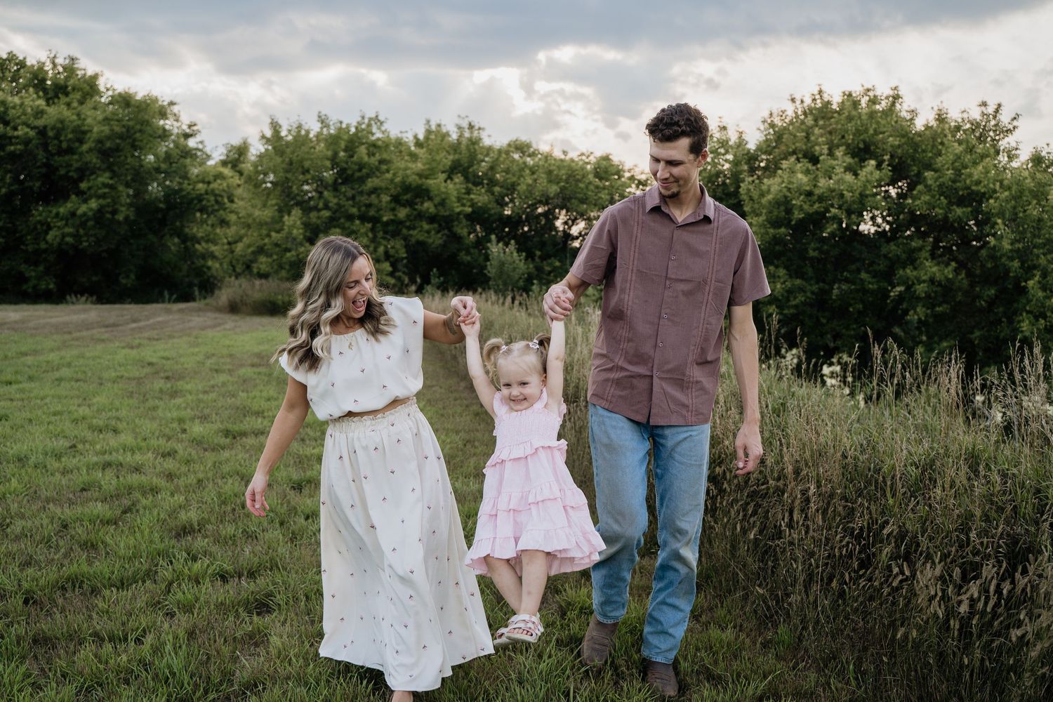 Happy young family walking through a lush green field at sunset, parents swinging their daughter between them.