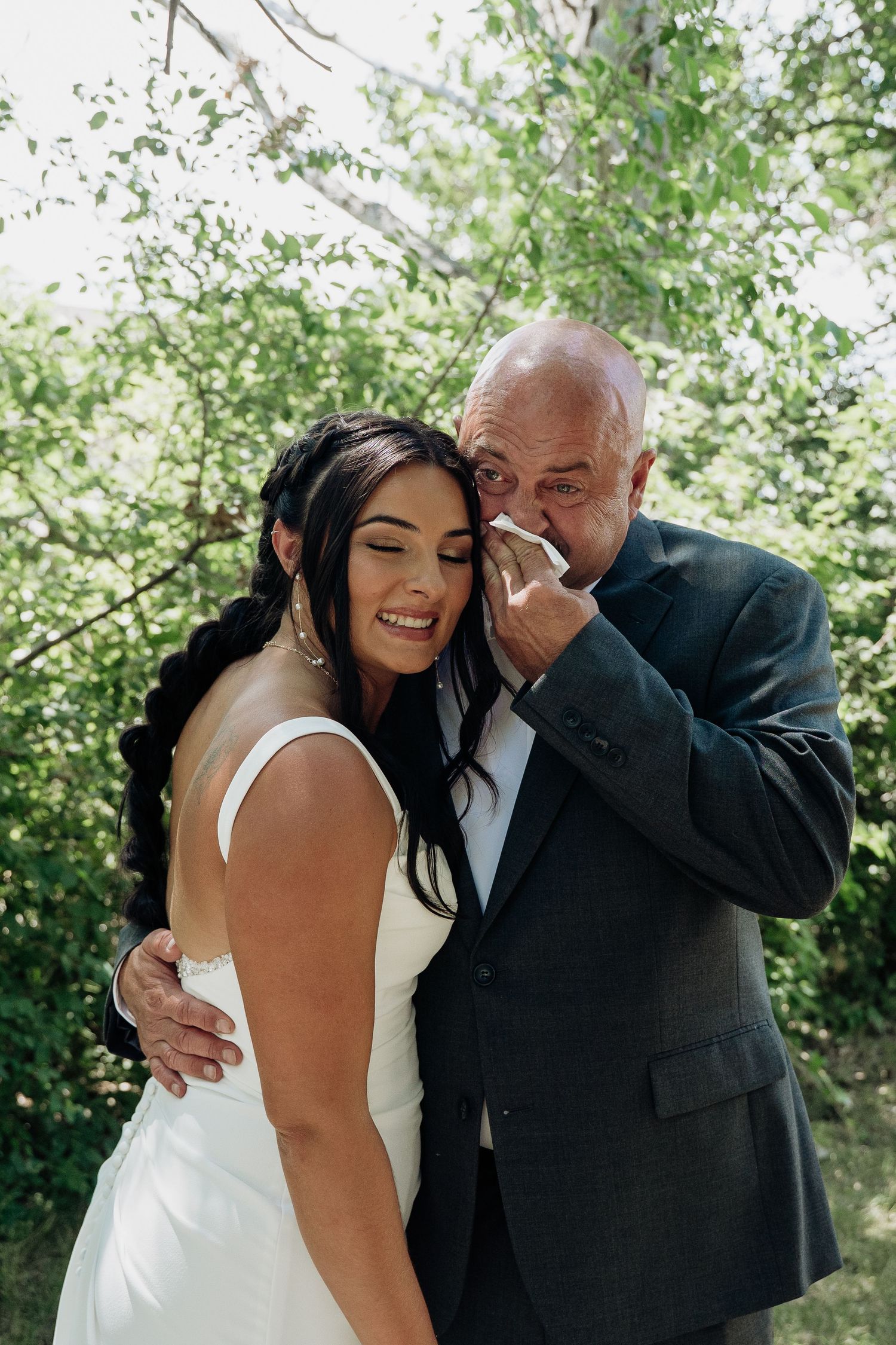 Emotional father wiping tears beside smiling bride in white dress outdoors among green trees.