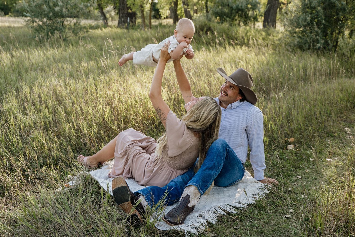 Young family sitting on blanket in green field, mother lifting baby in air while father watches lovingly.