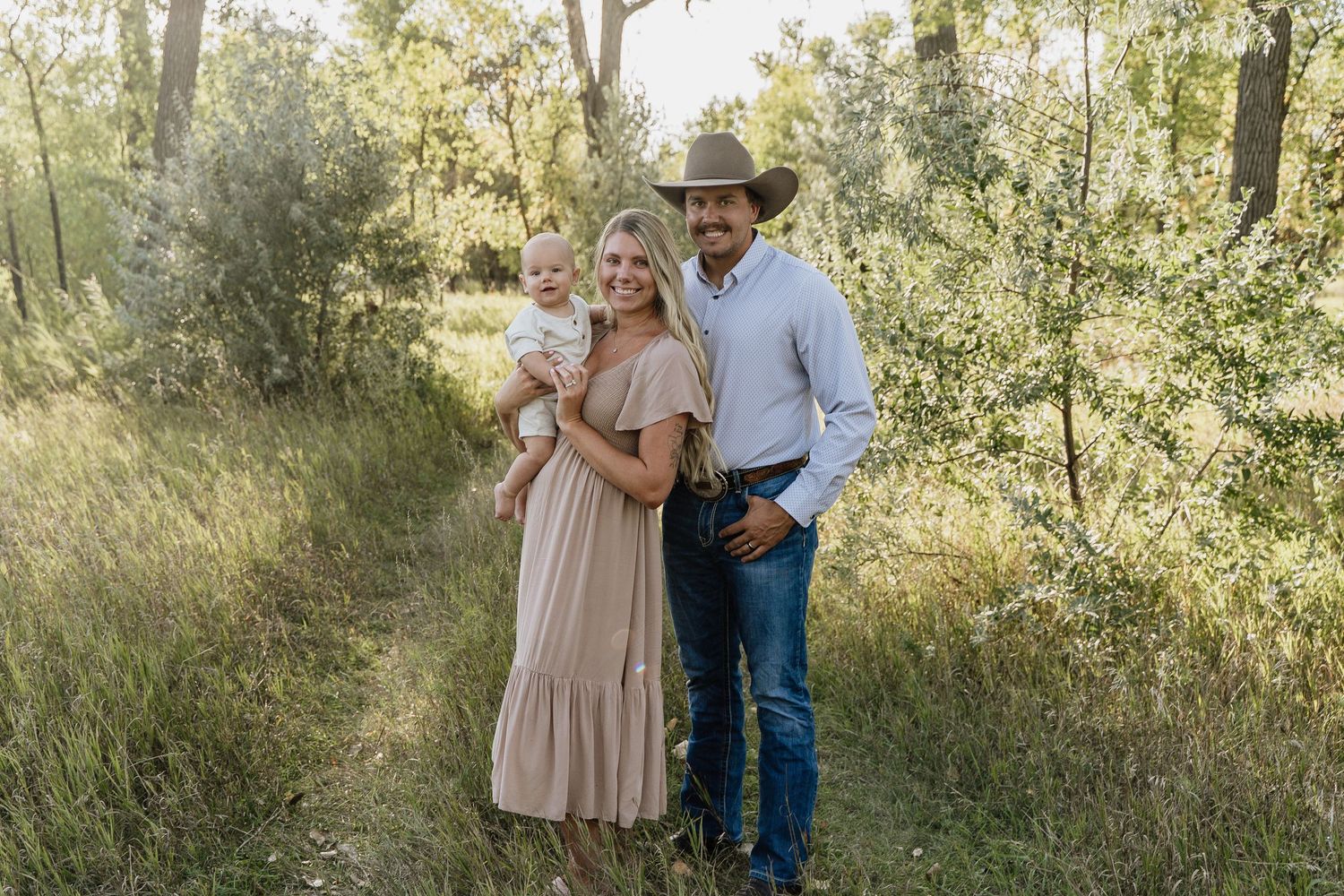 A young family poses on a wooded path, woman in pink dress holding baby, man in cowboy hat and jeans.