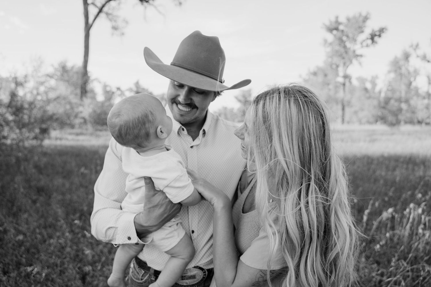 Black and white photo of smiling family with cowboy hat dad holding baby in outdoor field.