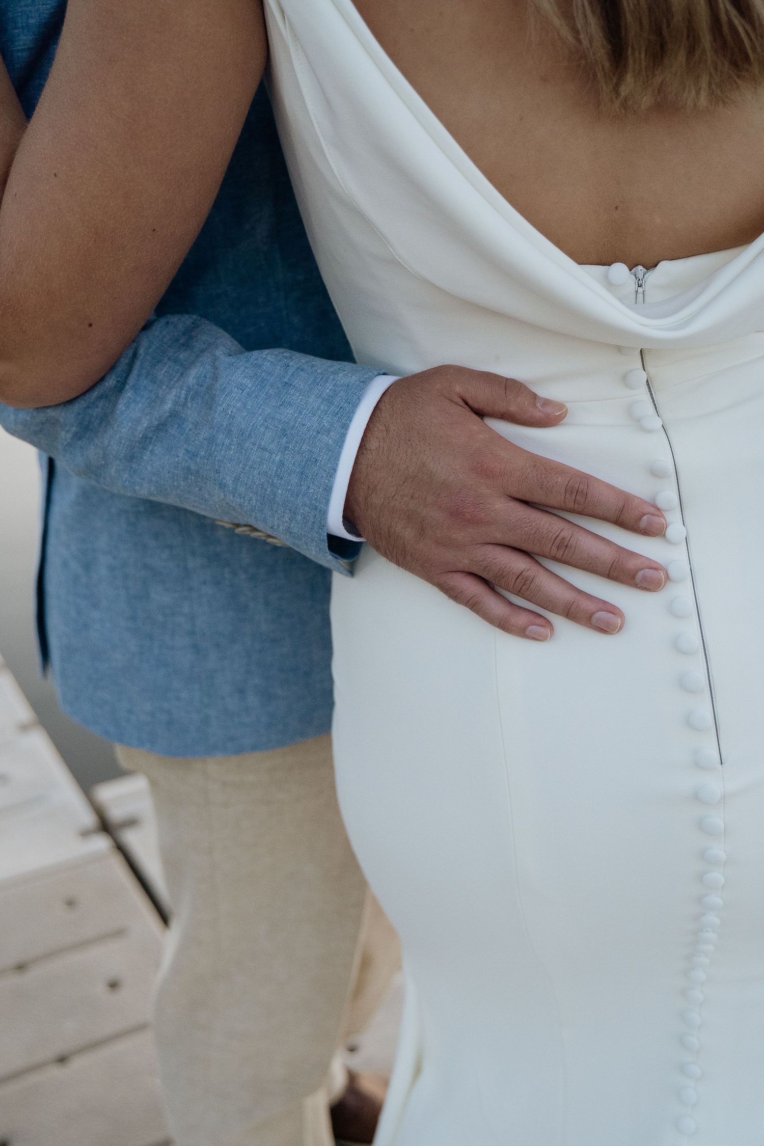 Close-up of groom's hand resting on bride's white button-back wedding gown at outdoor ceremony.