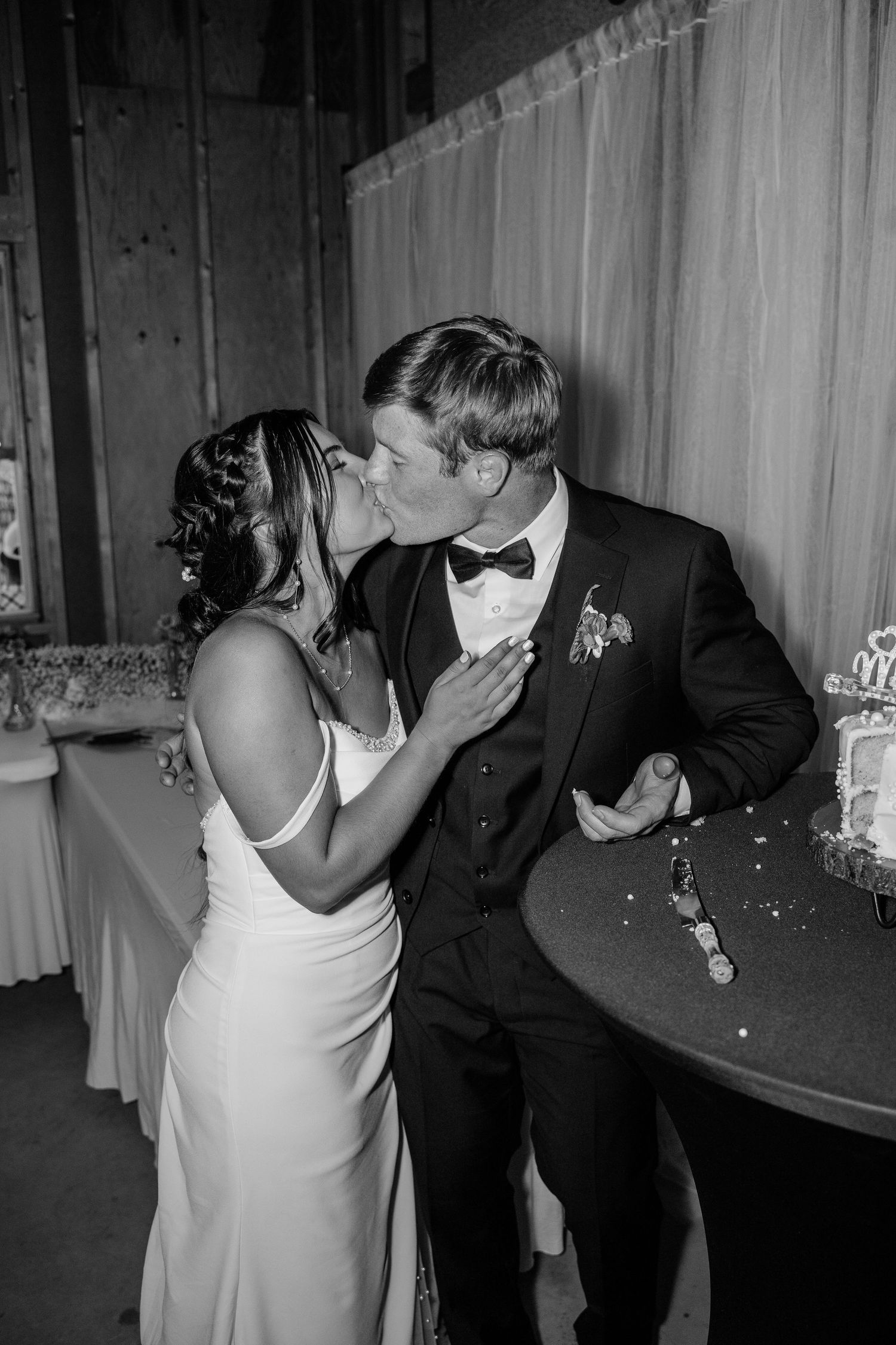 Black and white photo of bride kissing groom on the cheek near wedding cake at reception.