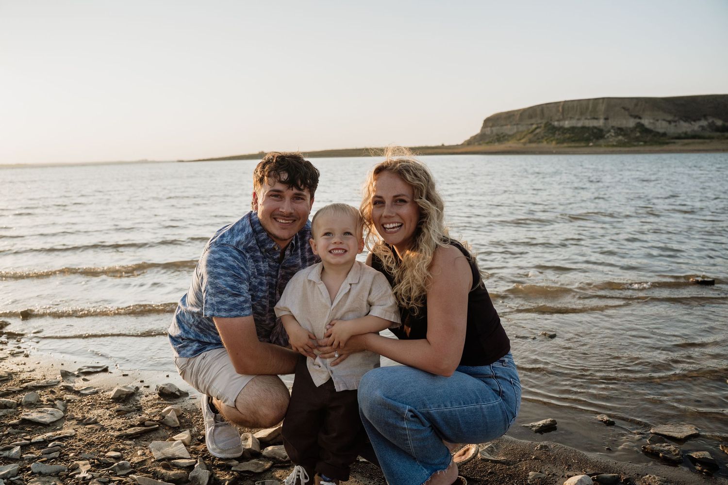 Happy young family with a baby posing on a rocky beach with a mesa in the background at golden hour.