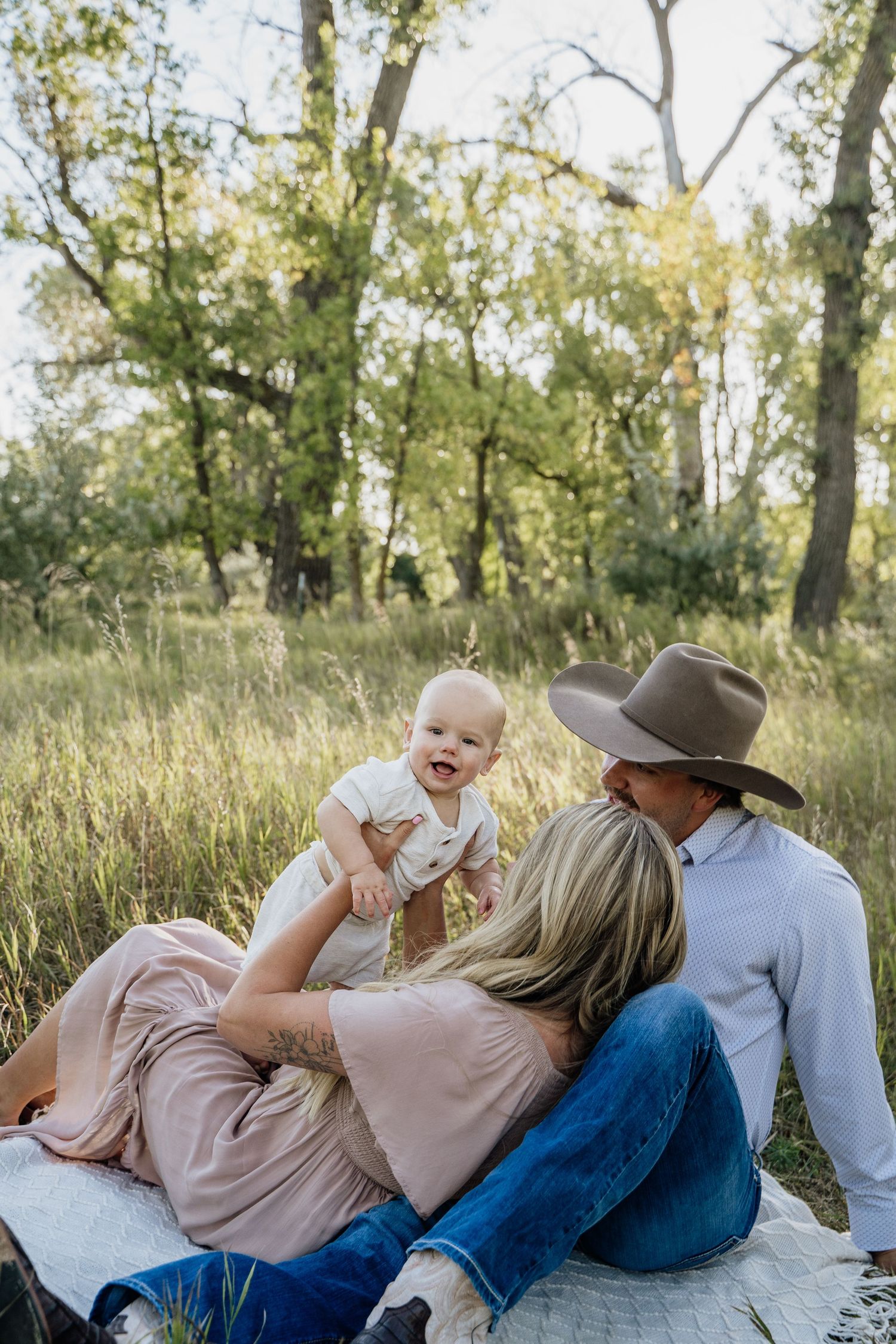 Young family sitting in green meadow, parents holding laughing baby outdoors.