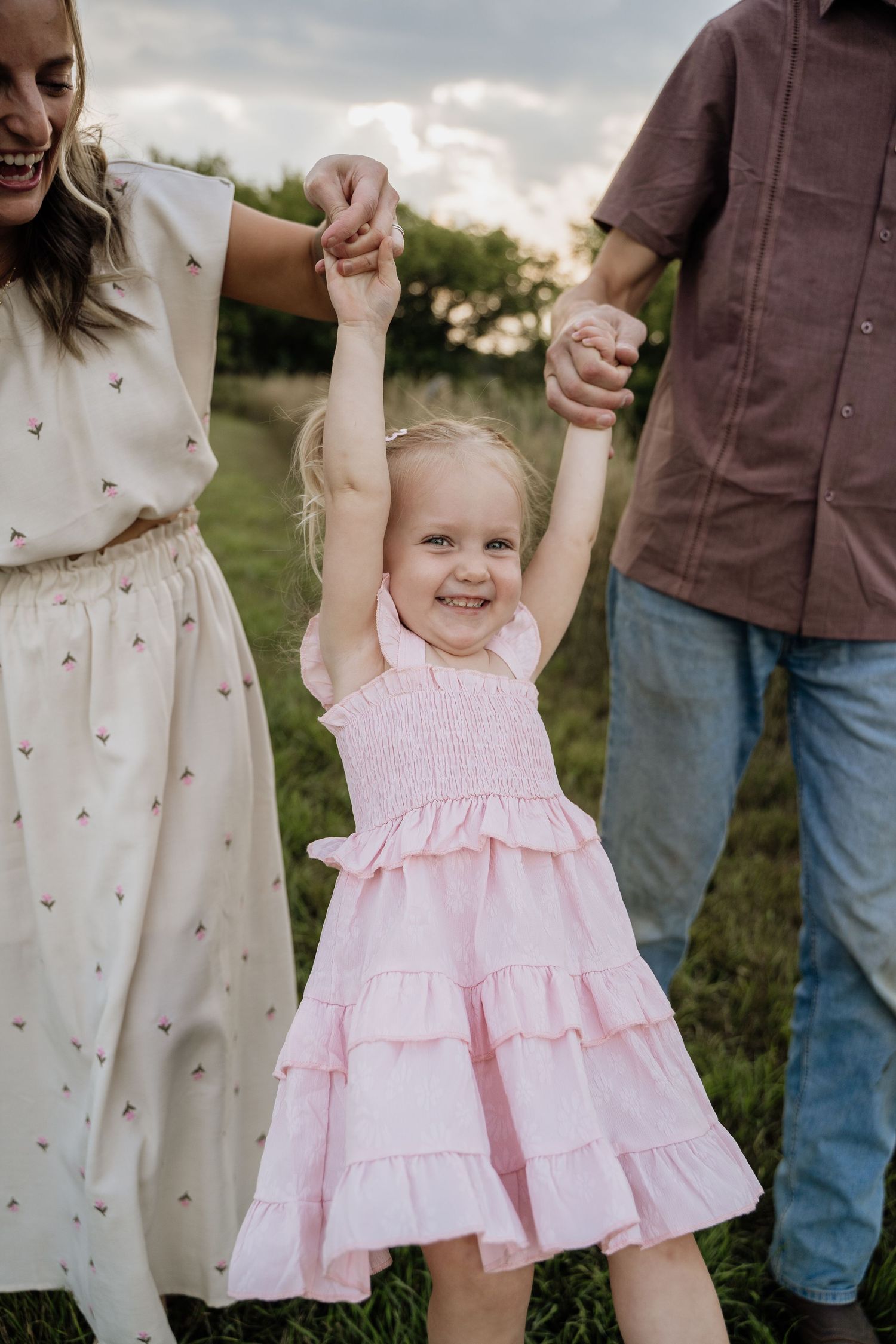 Joyful toddler girl in a pink dress raising her arms while held by parents in a green field at dusk.