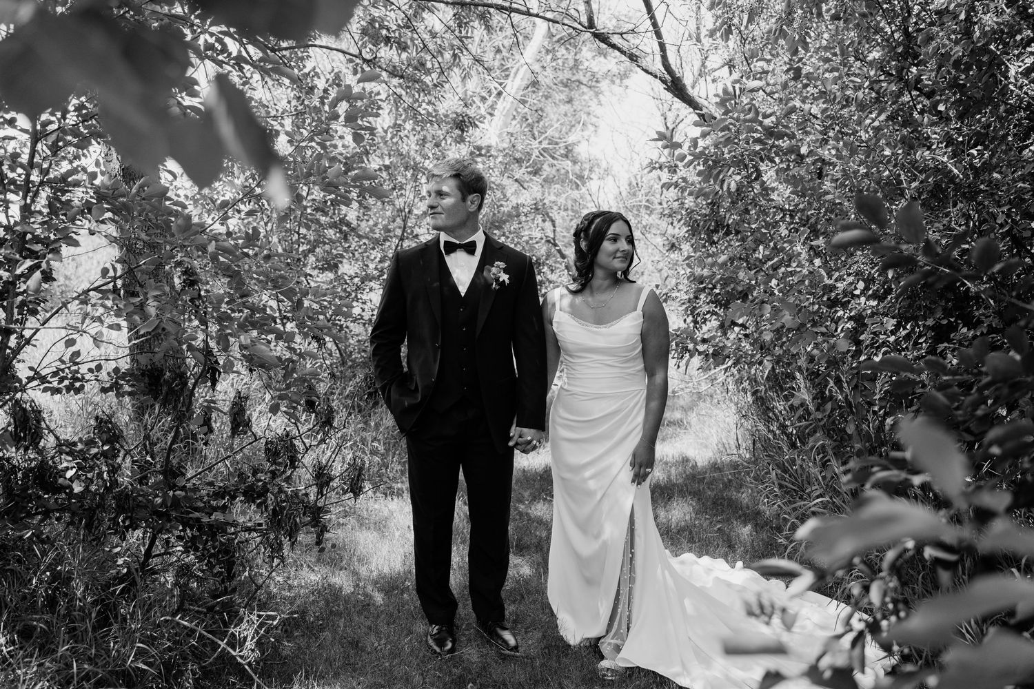 Black and white photo of a bride and groom holding hands while walking through lush green trees on their wedding day.