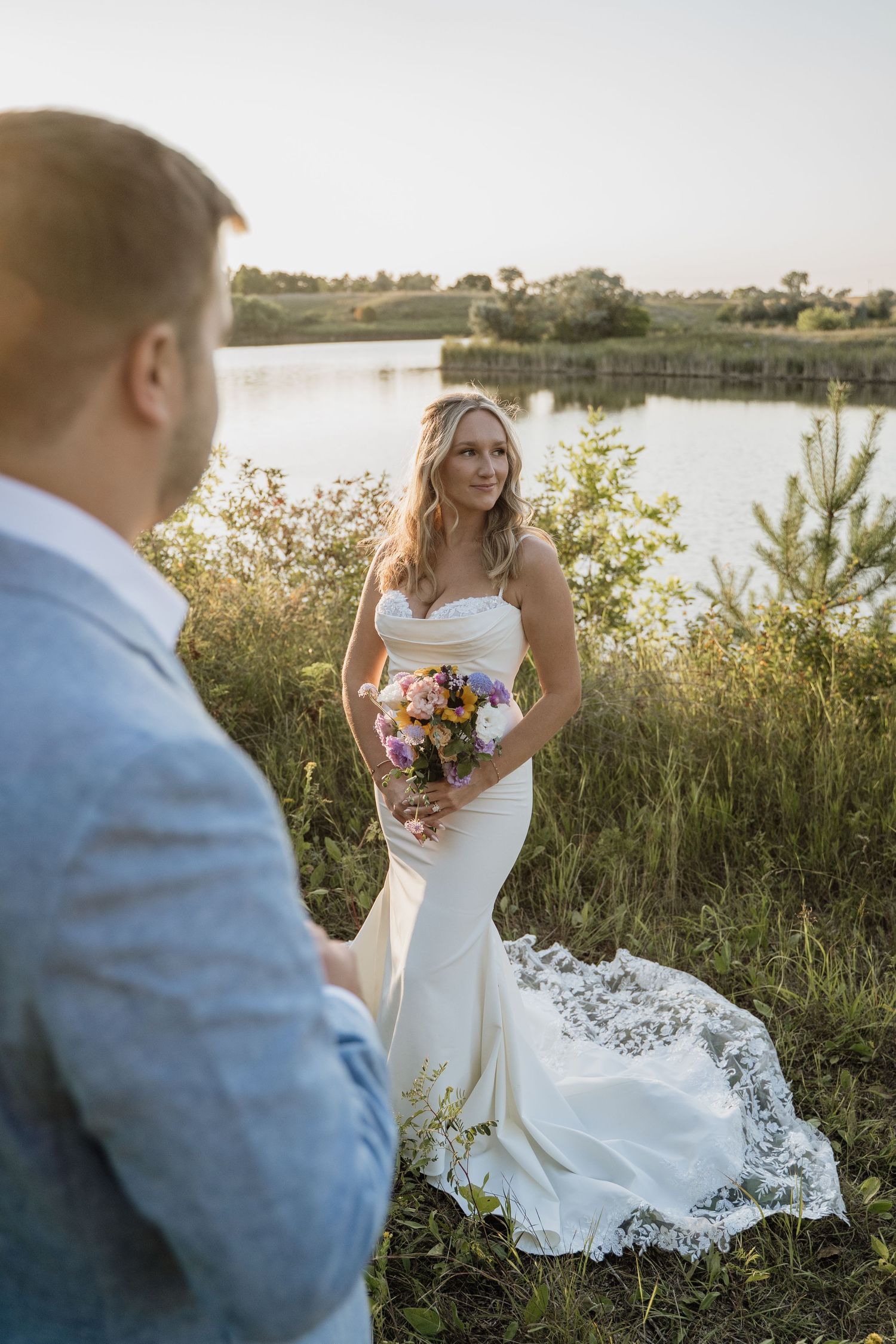 Bride in a mermaid gown holding a colorful bouquet smiling at groom near a sunlit lake.
