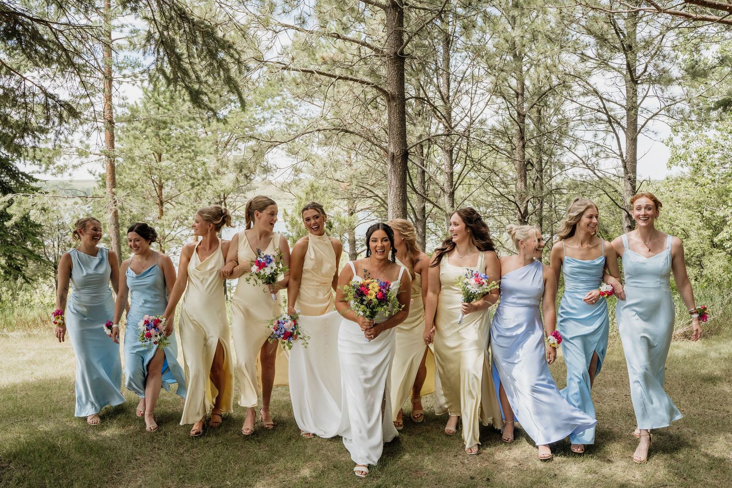 Bride and full bridal party in cream and blue dresses walking through a sunny wooded area.