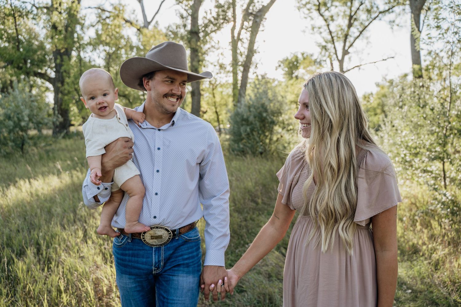 Smiling western family with man in cowboy hat holding baby while walking hand in hand outdoors.