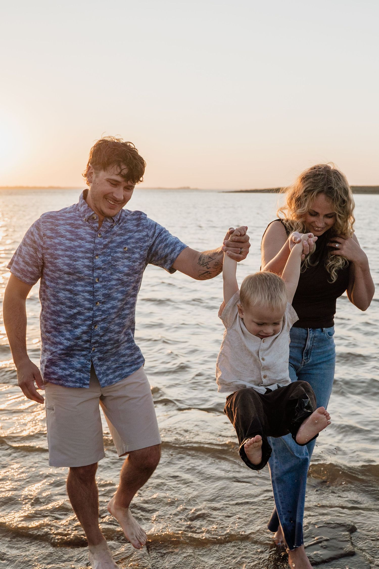 Joyful parents swing their laughing toddler between them while walking along a sunlit lakeshore at sunset.