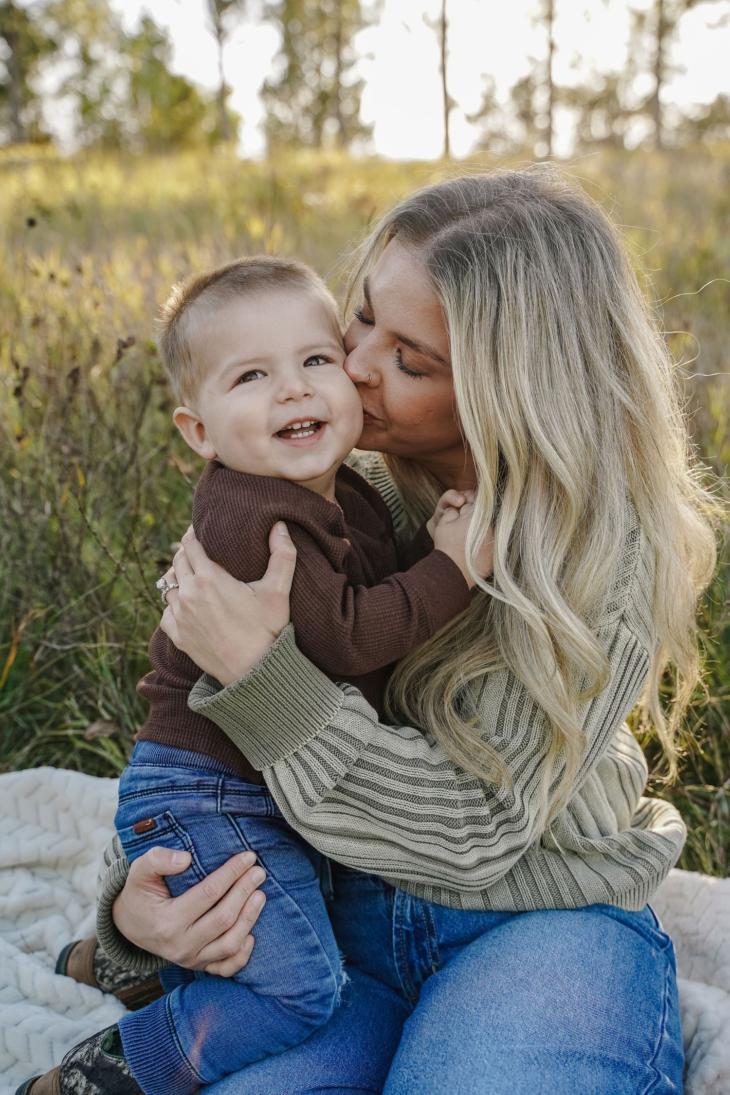 Mother kissing smiling baby boy on cheek outdoors in a sunlit field.