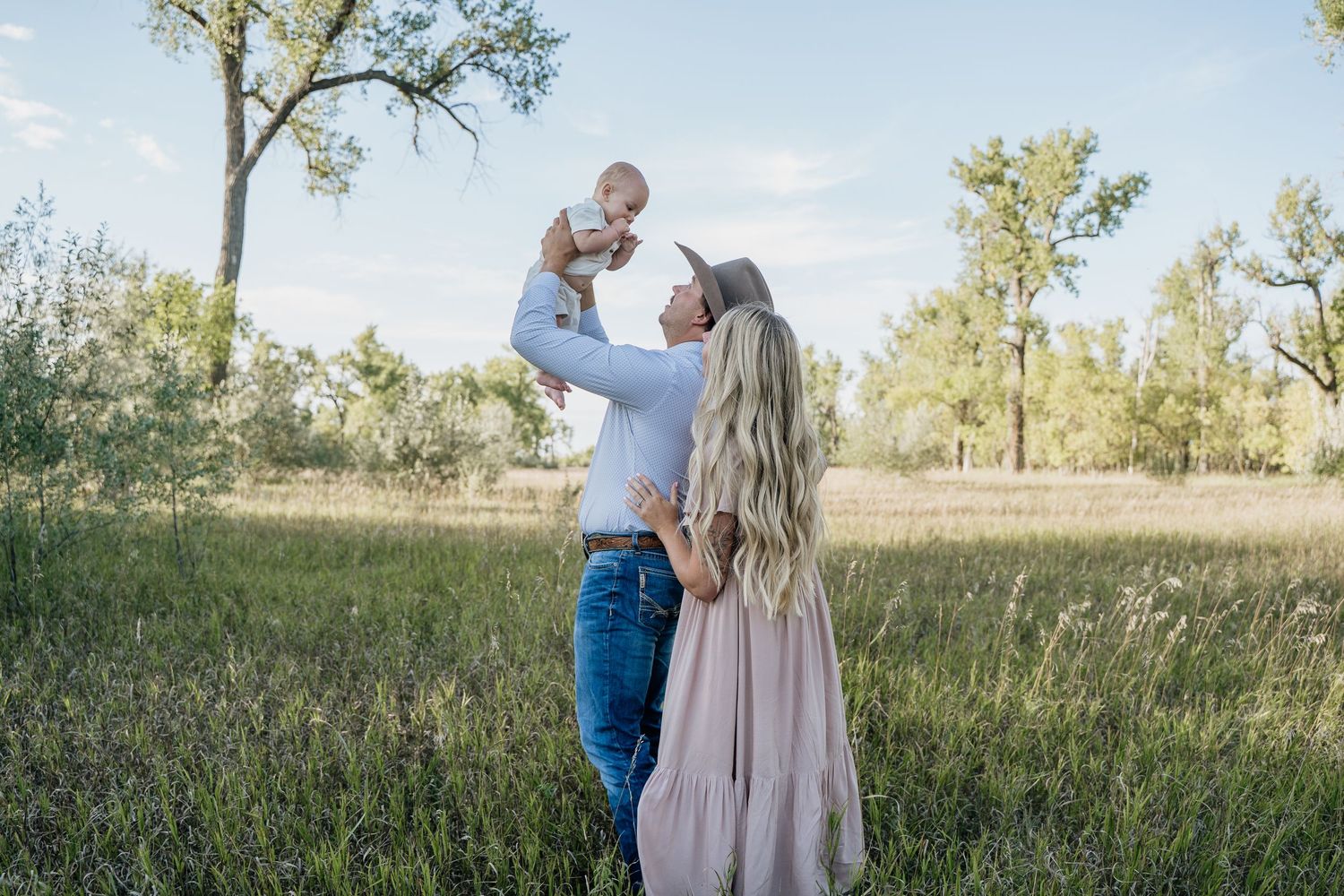 Parents lifting laughing baby in the air in a sunny green meadow with trees in background.