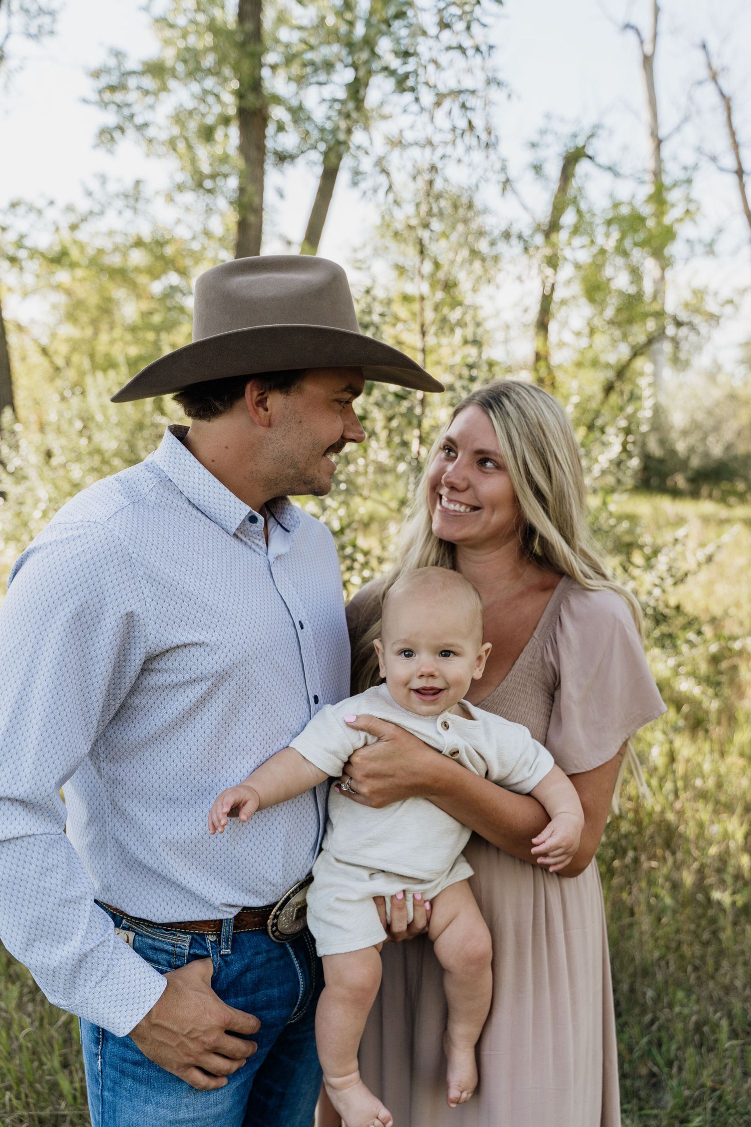 Smiling couple in rustic western style holding baby outdoors in green wooded setting.