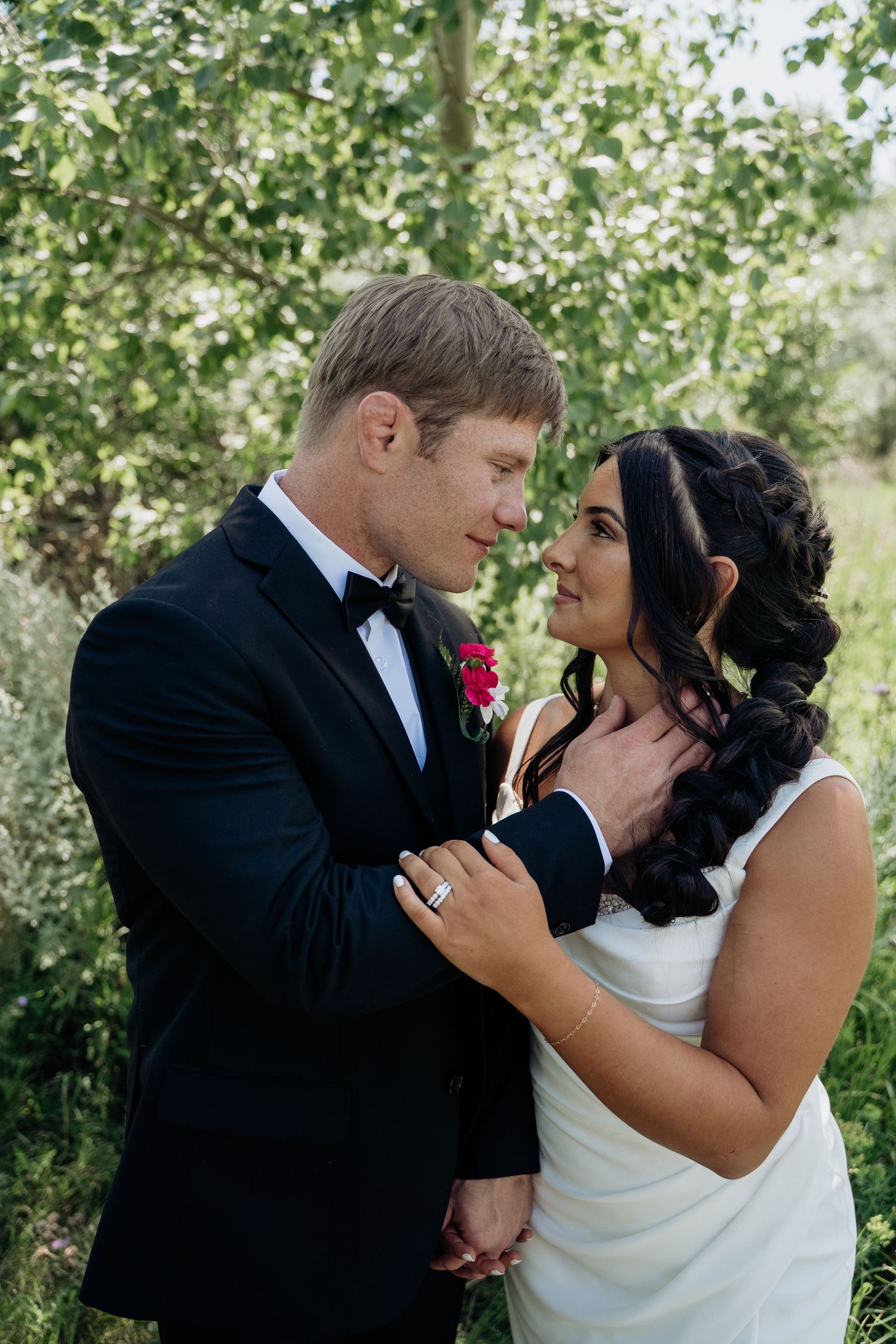 Bride and groom gaze into each others eyes while embracing in front of green trees.