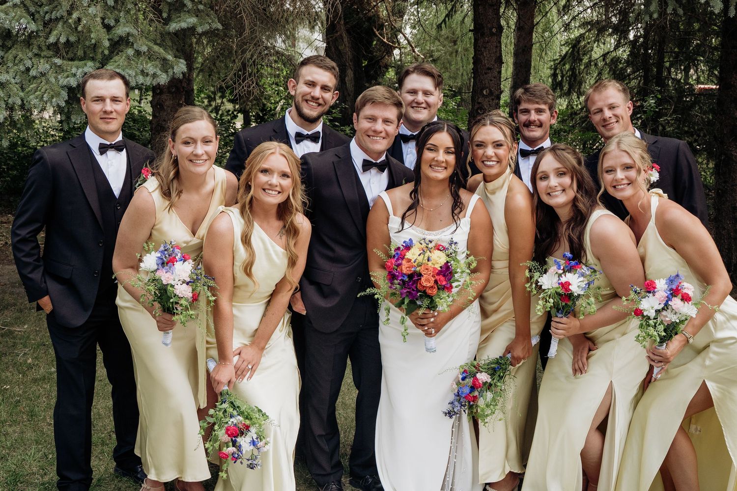 Bride and groom surrounded by smiling wedding party in cream dresses and black tuxedos outdoors.