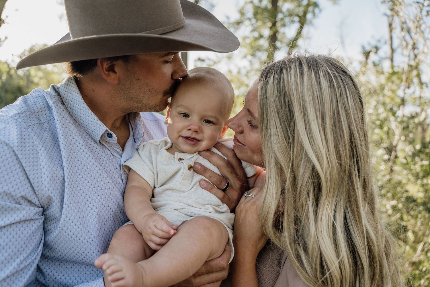 A couple in a natural outdoor setting kissing their smiling baby, with the man wearing a cowboy hat.