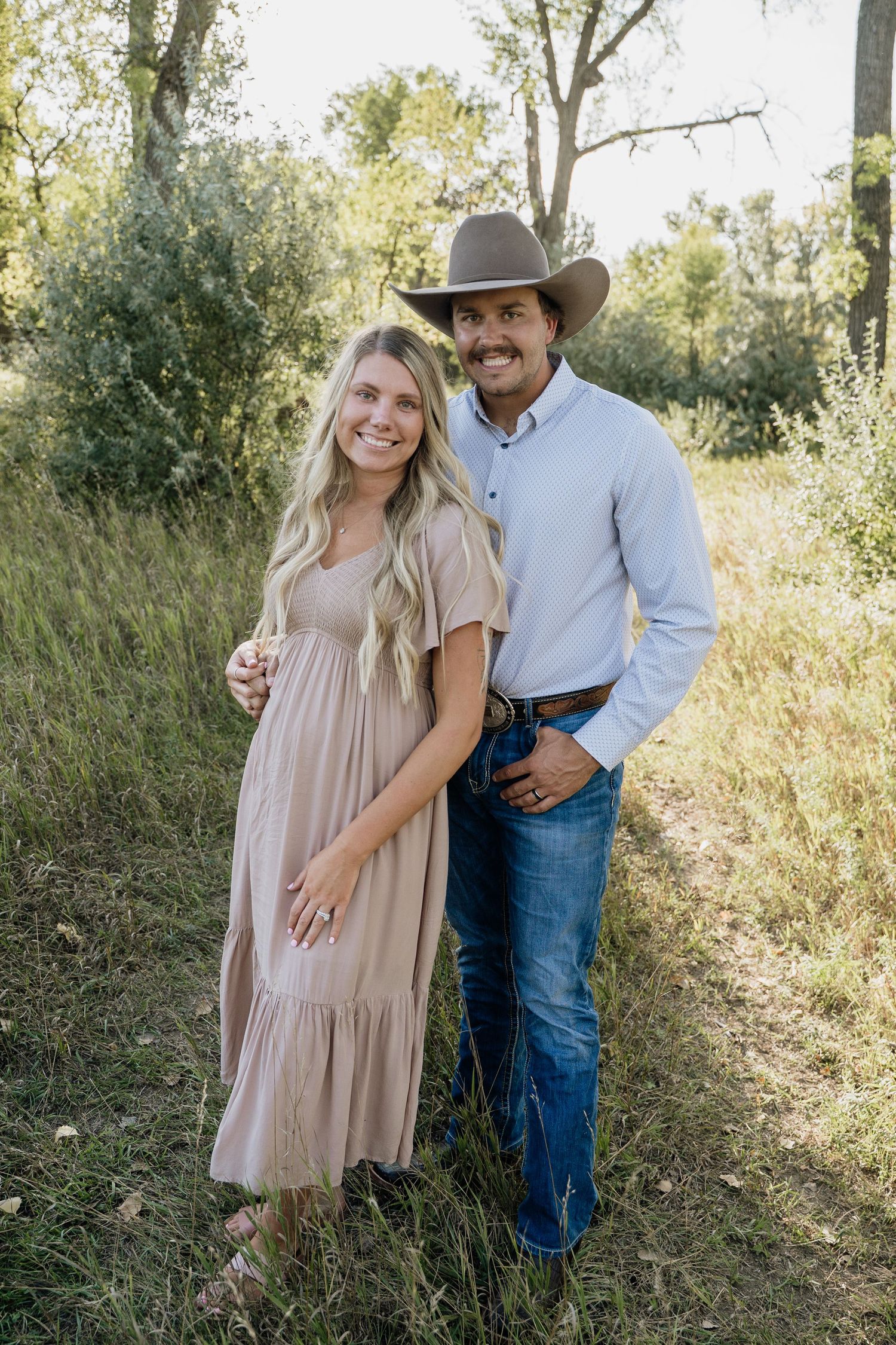 Smiling couple in field, woman in beige dress and man in cowboy hat and jeans.