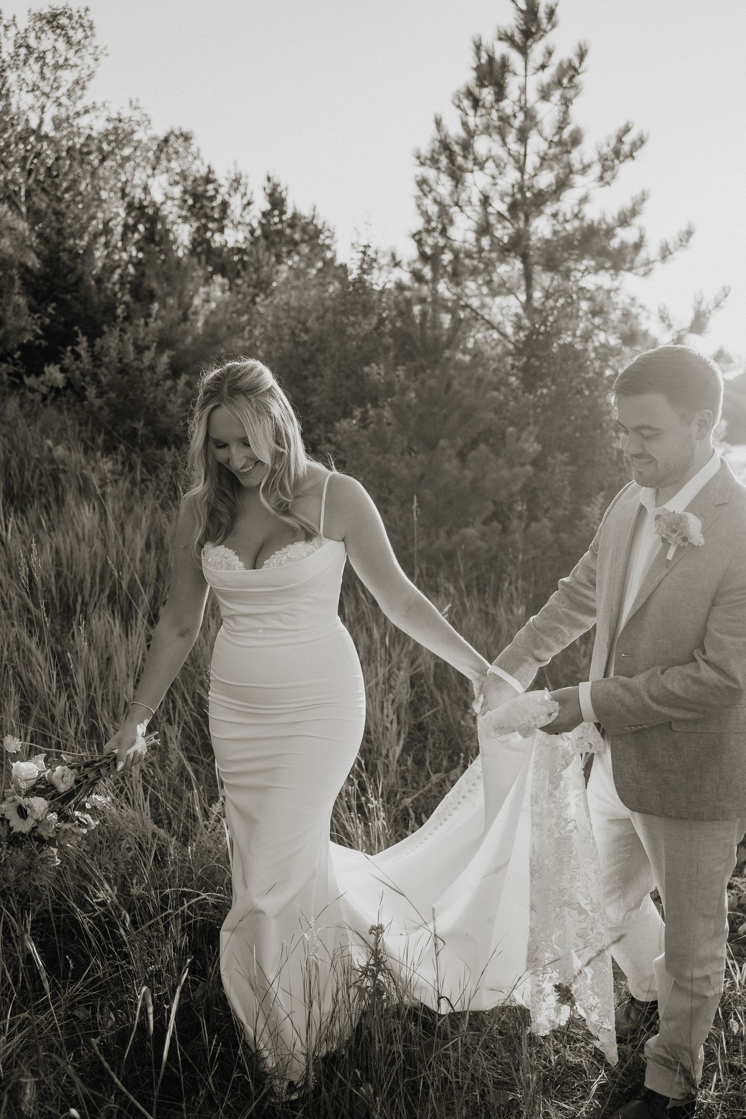 Black and white photo of groom helping bride with her train while walking through a field.