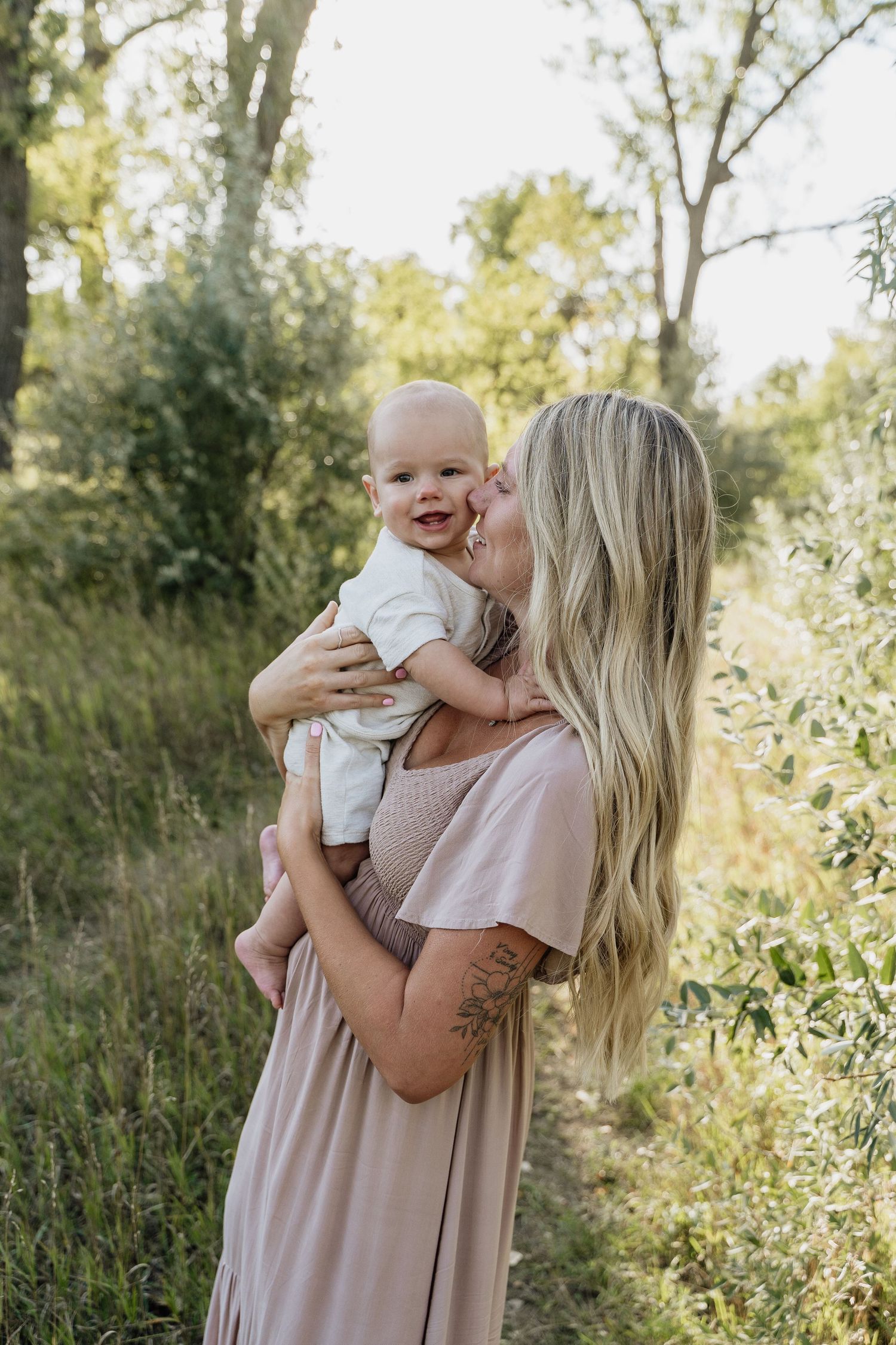 Blonde woman in blush maxi dress kissing smiling baby outdoors in lush green nature setting.