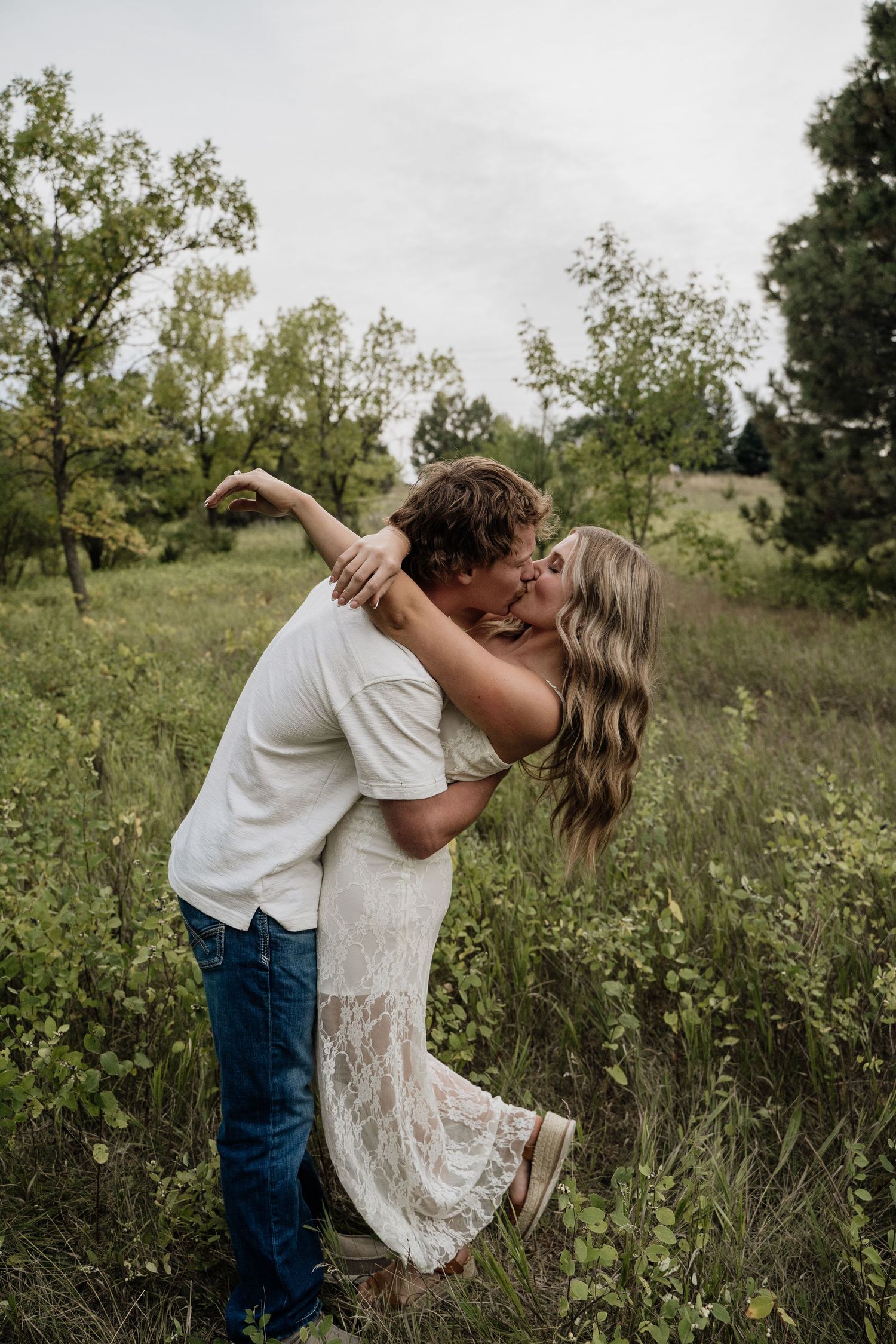 Couple sharing romantic dip kiss in wildflower field, man in white shirt and jeans, lace dress.