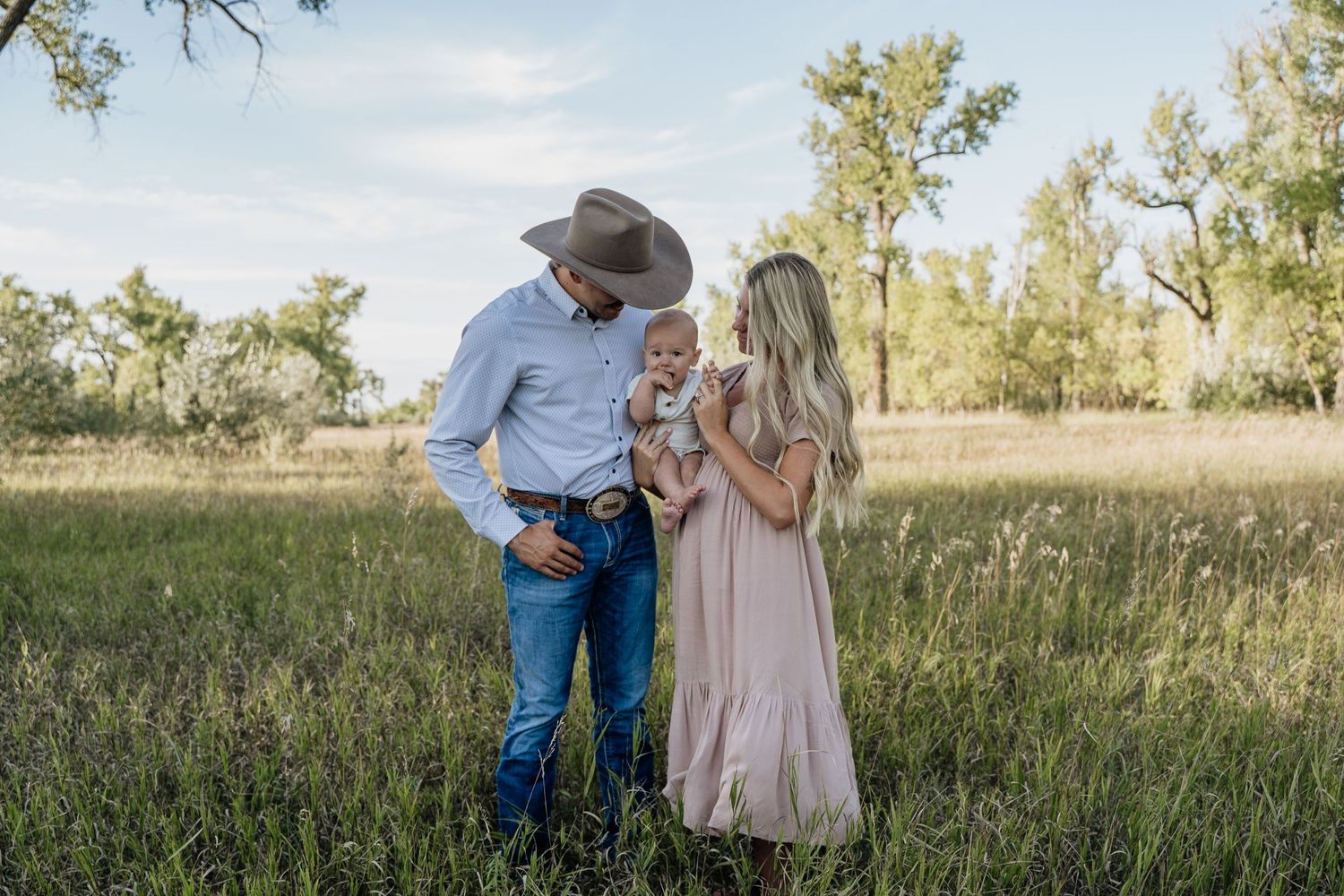 A young family with a cowboy hat-wearing father, mother in pink dress, and baby stand in a lush green meadow.