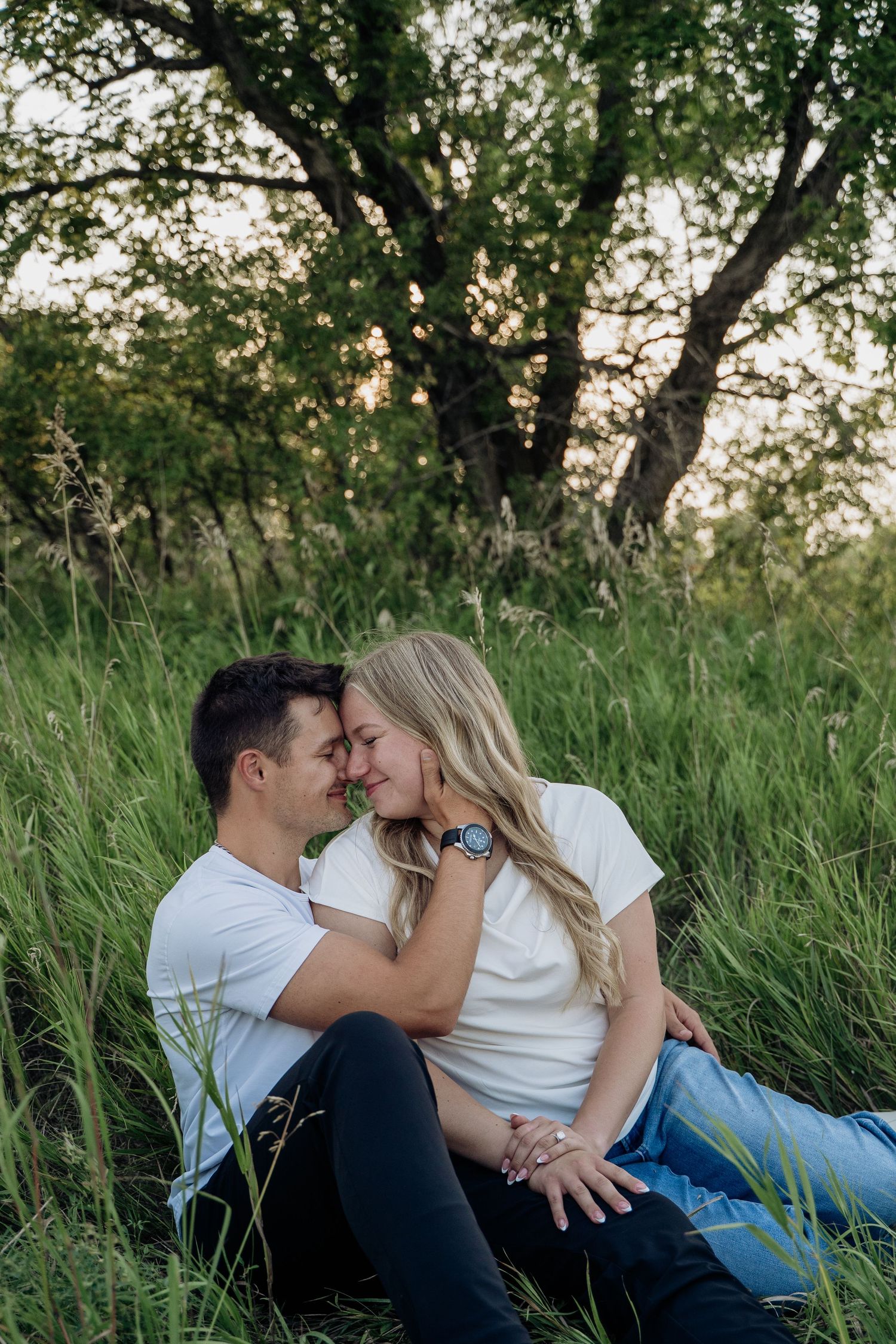A romantic couple sits in tall green grass, touching foreheads and smiling at sunset.