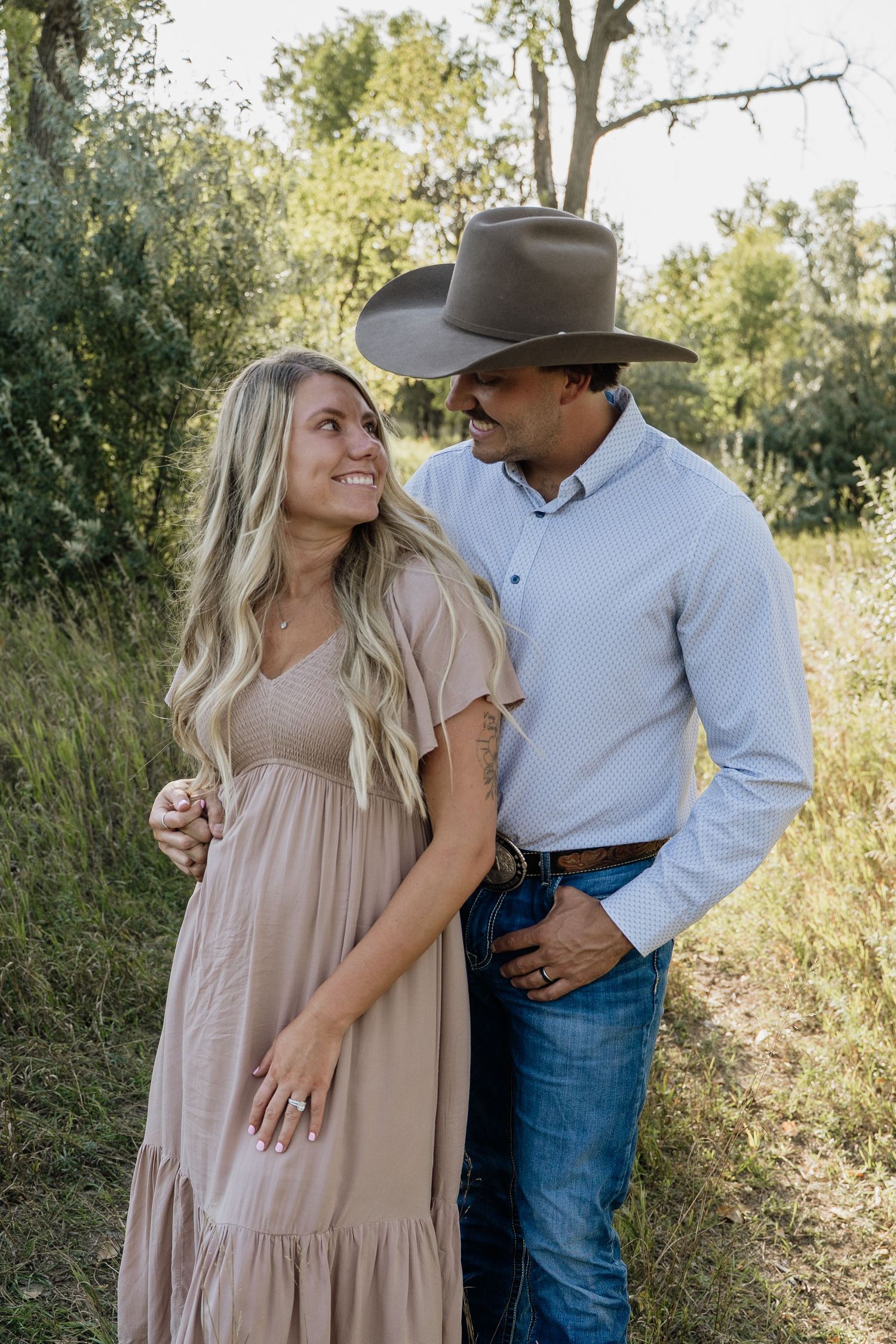 Smiling couple in rustic outdoor setting, woman in blush dress and man in cowboy hat and jeans.