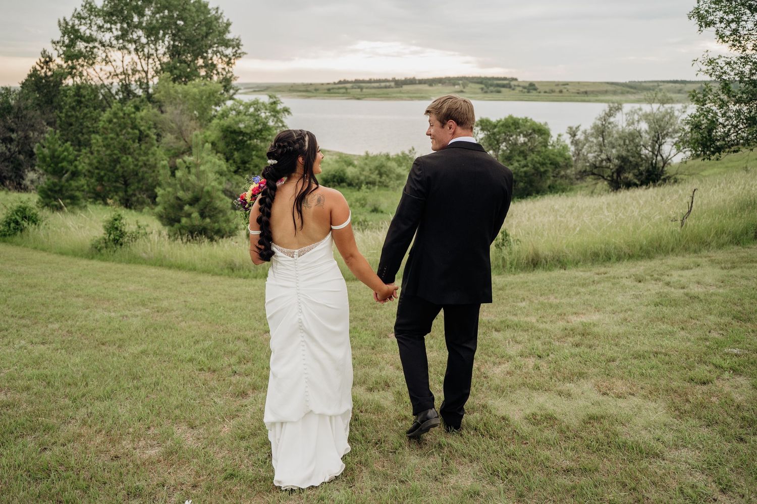 Bride and groom holding hands walking toward a scenic lake surrounded by lush green hills at dusk.