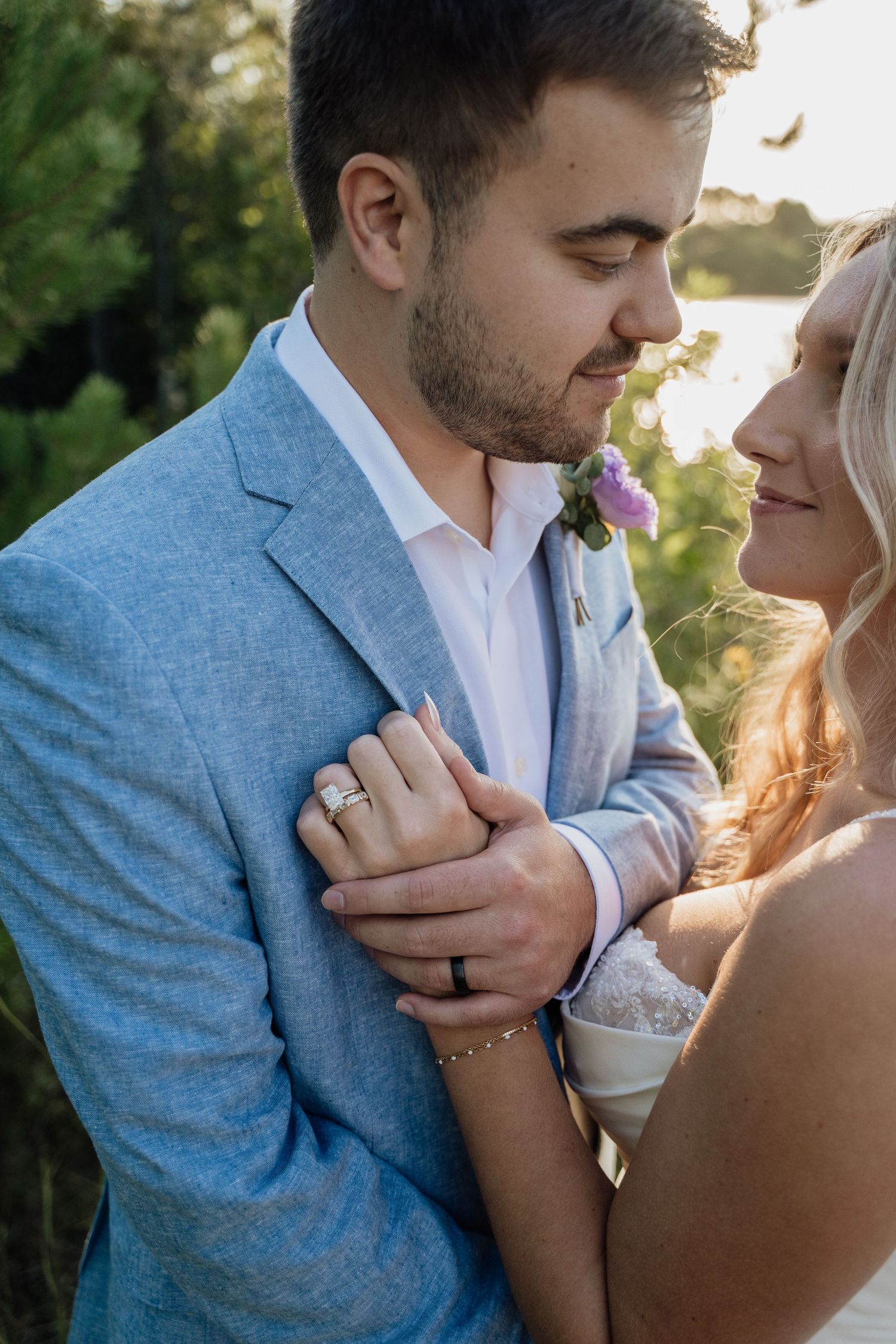 Bride embraces groom in blue suit during golden hour outdoor wedding portrait near waterfront.