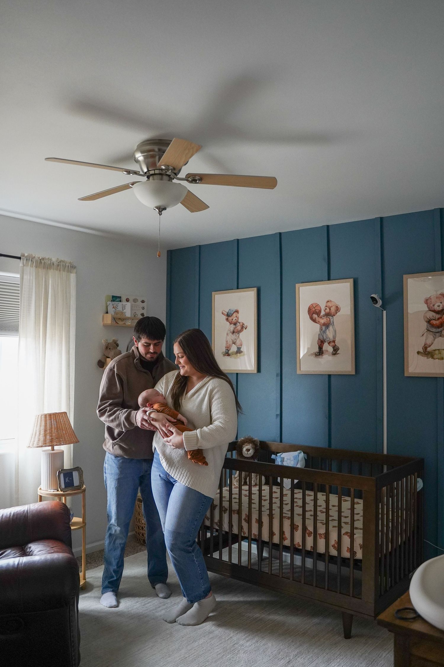 New parents holding newborn baby in blue nursery with crib and vintage sports artwork.