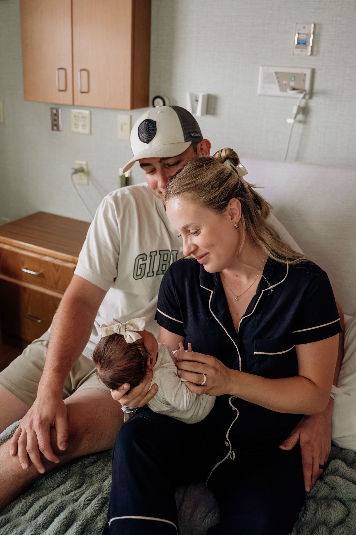 New parents in hospital room admiring their newborn baby, father wearing a baseball cap.
