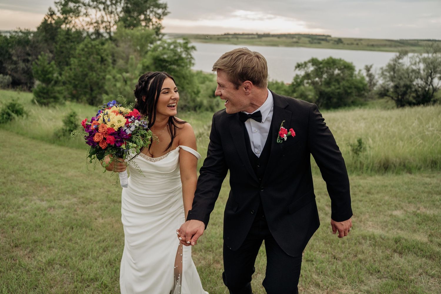 Bride and groom laughing together holding hands in a lush green meadow with lake views.