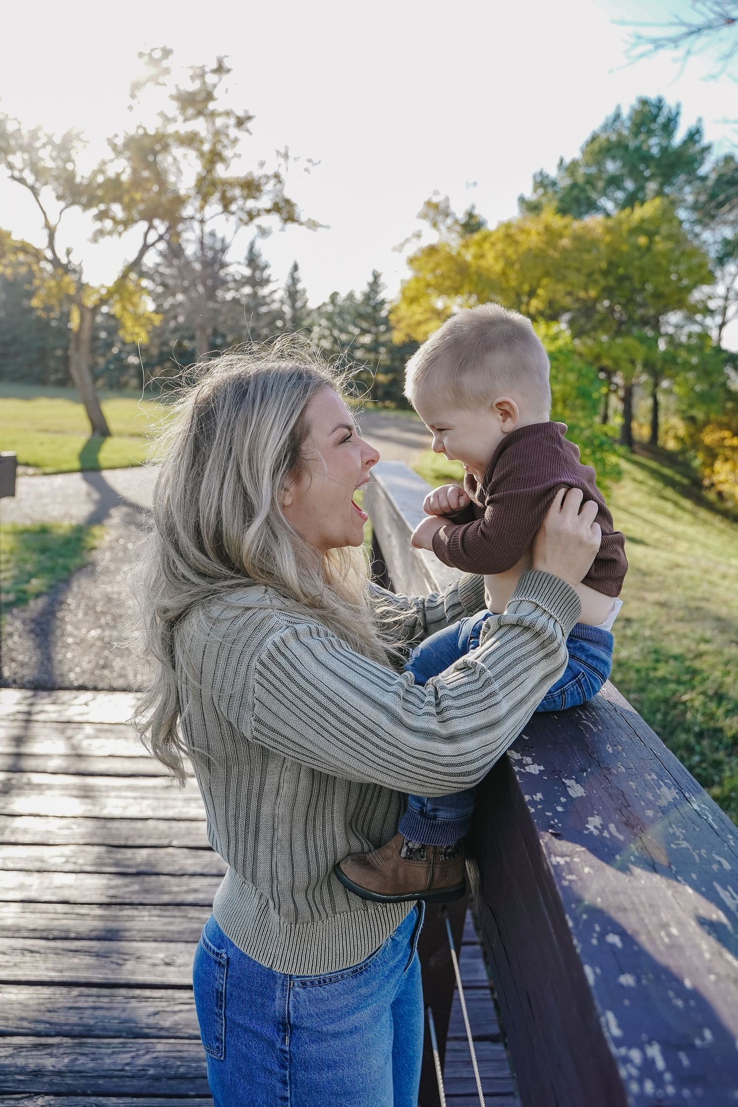 Blonde mother holding laughing baby on a wooden bridge in a sunny park.