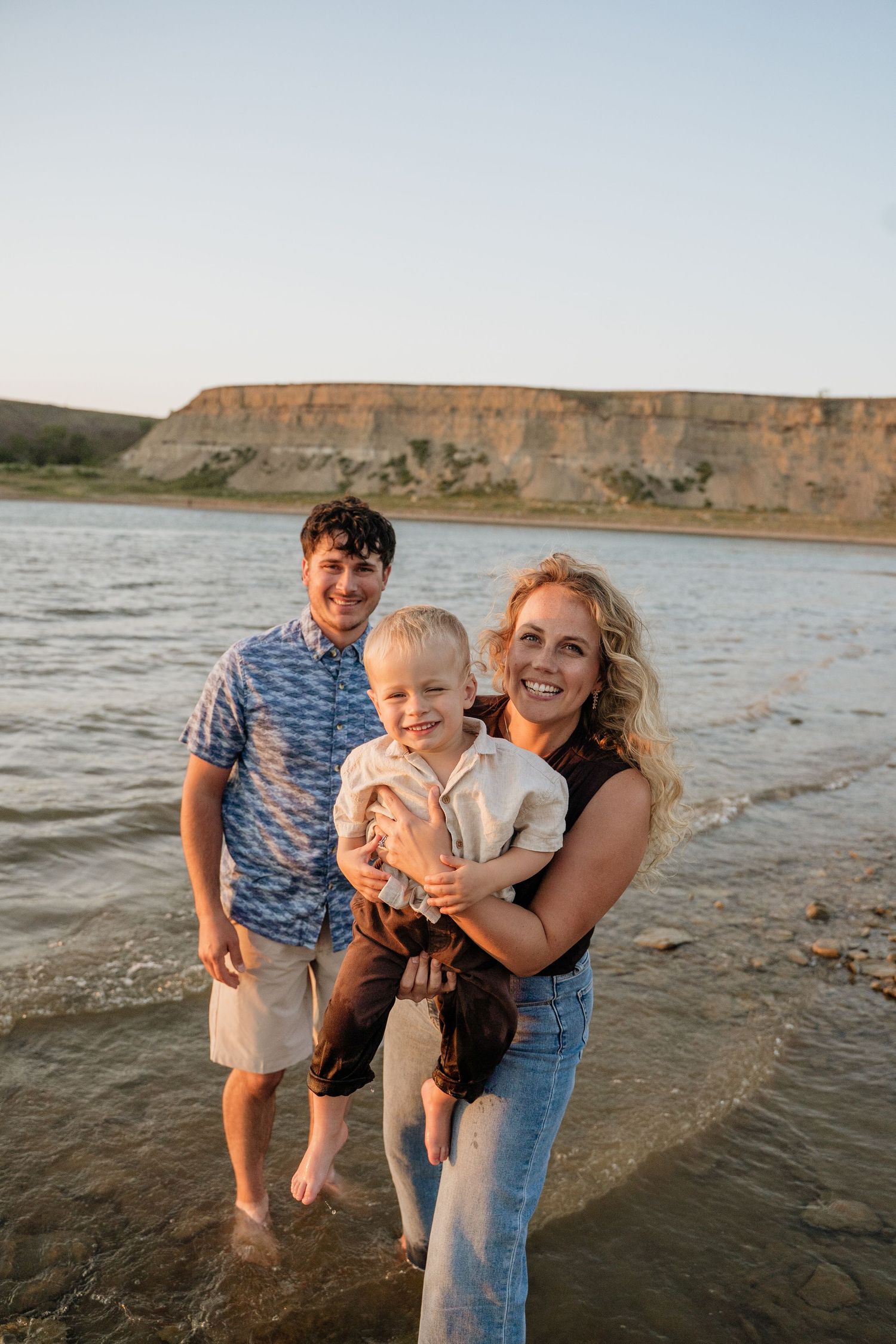 Smiling family of three stands in shallow water with scenic cliffs and golden sunset sky behind them.