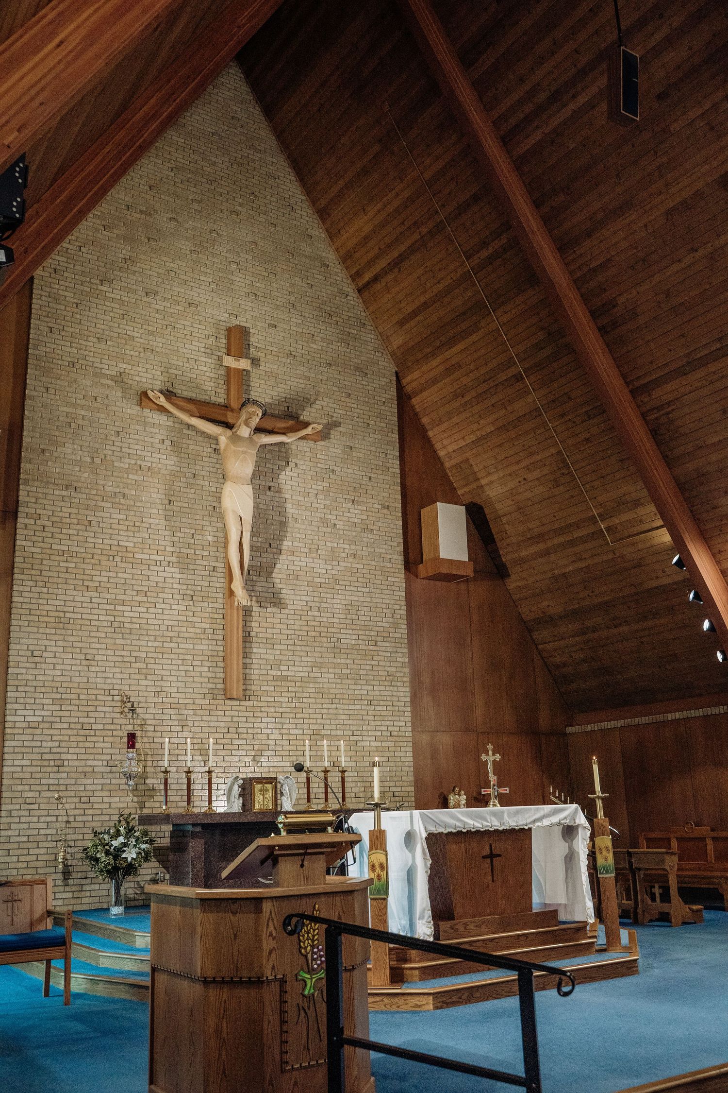 Church altar with crucifix on brick wall, wooden ceiling, candles, and blue carpet.