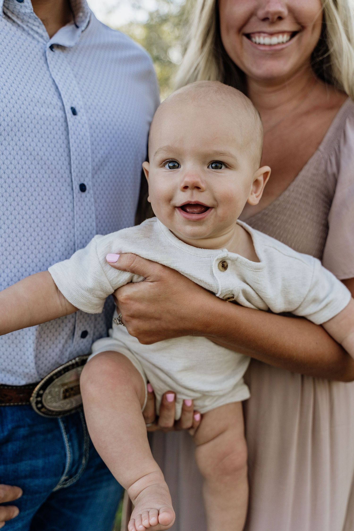 Smiling baby in cream onesie held by parents outdoors during a family photo session.