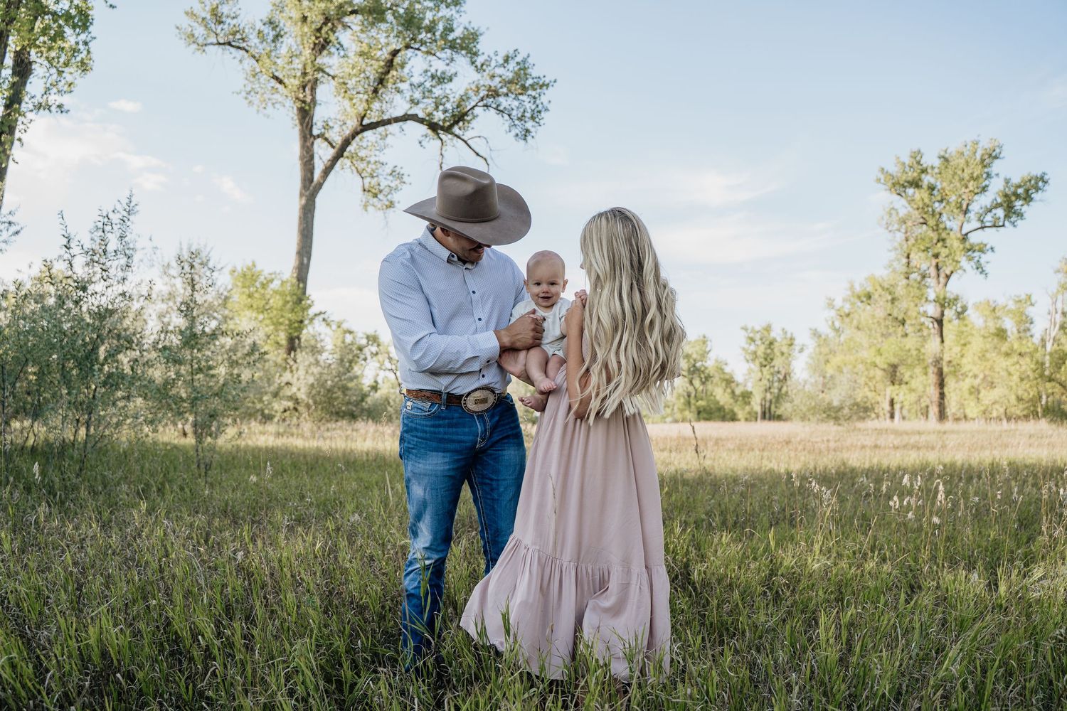 Couple in field, man in cowboy hat and woman in pink dress holding newborn baby.