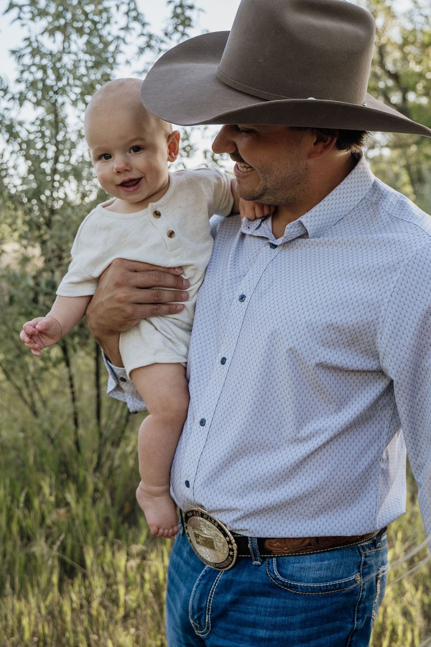 Proud father in cowboy hat and blue shirt holding smiling baby boy outdoors in nature.