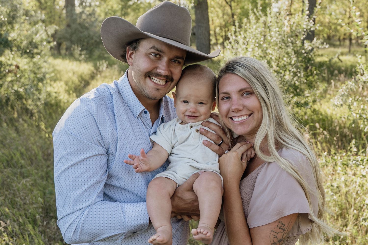 Smiling couple with cowboy hat holding happy baby outdoors in green nature setting.