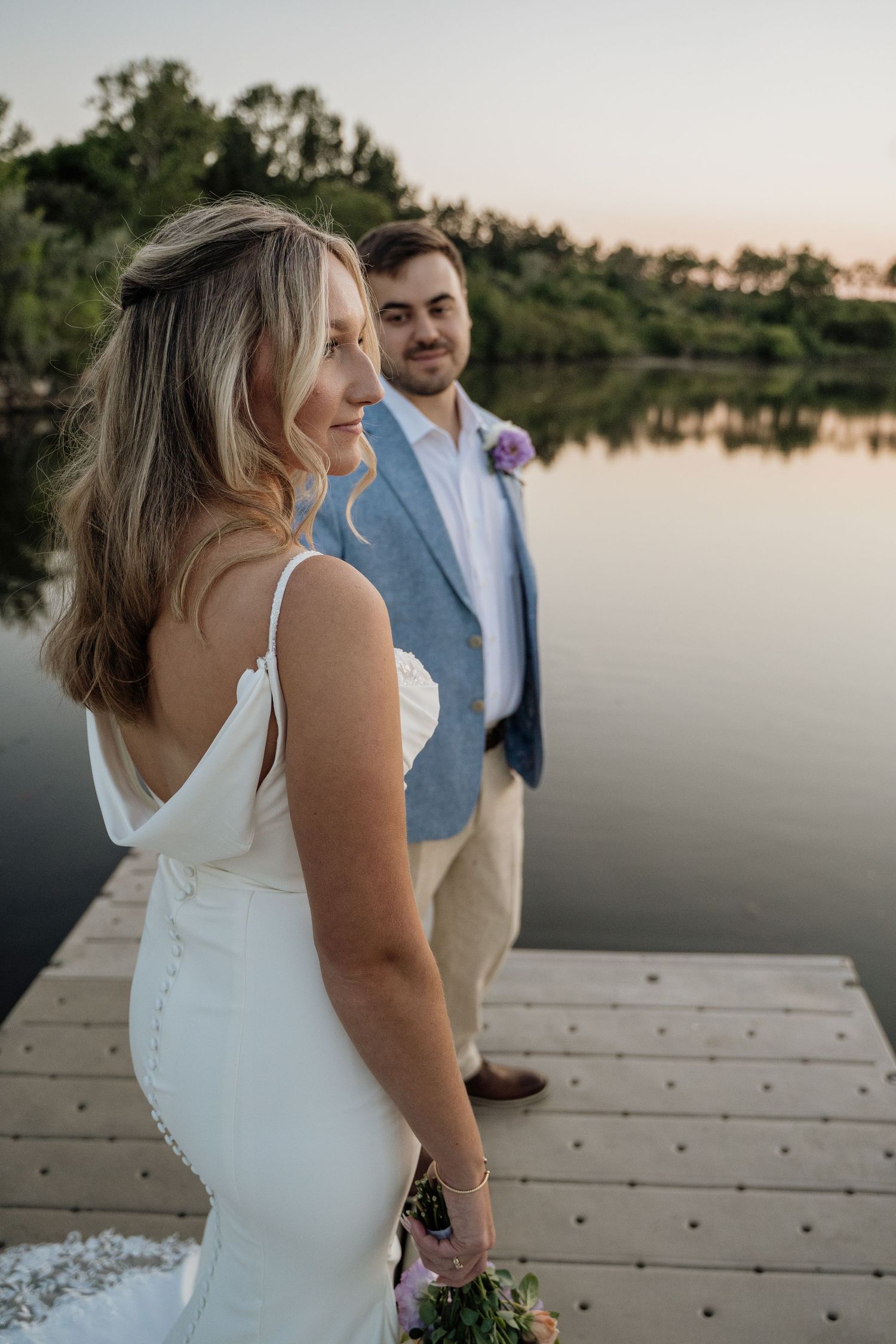 Bride in white gown smiles on a wooden dock as groom in blue blazer stands behind her at sunset.