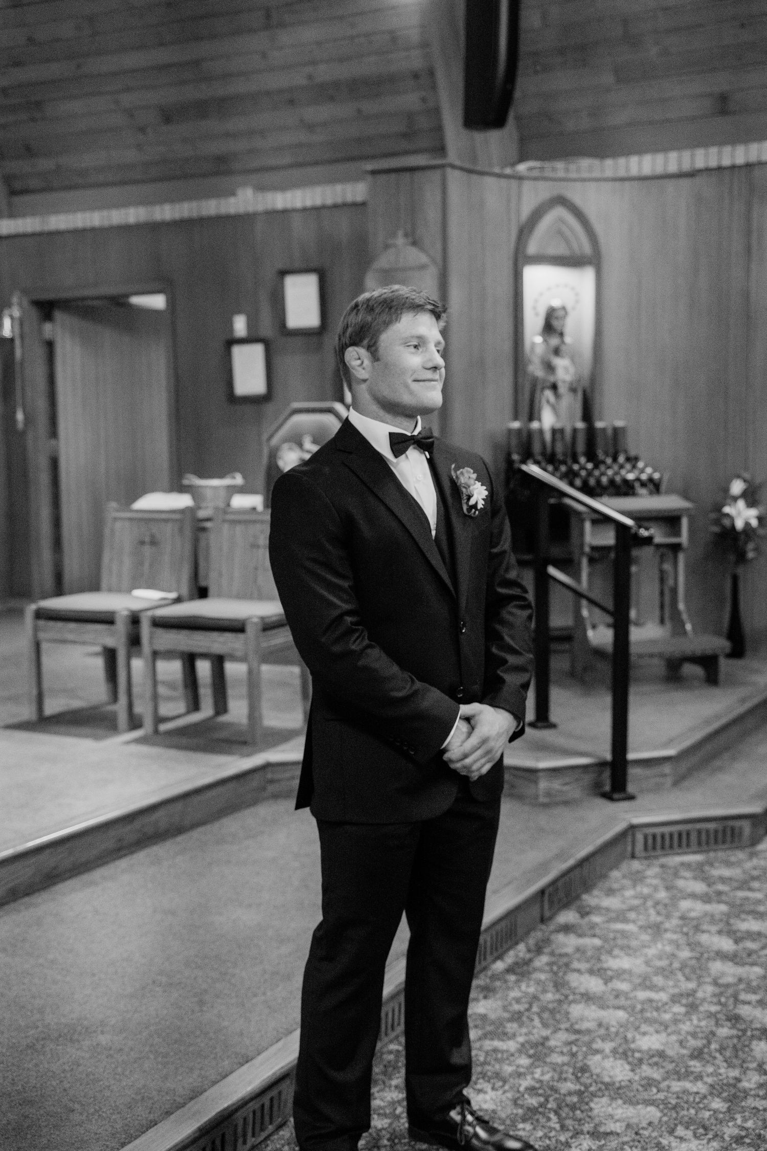 Groom in black tuxedo smiling at church altar during wedding ceremony, black and white photo.