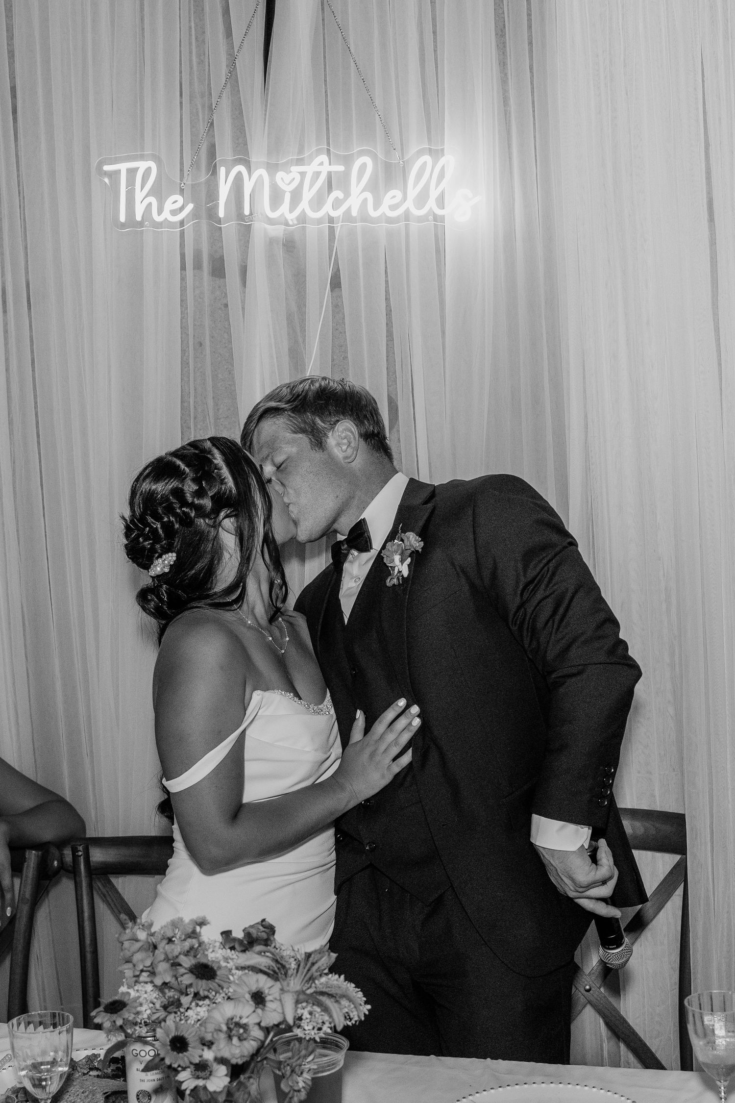 Bride and groom share a kiss beneath a neon sign reading The Mitchells at wedding reception.