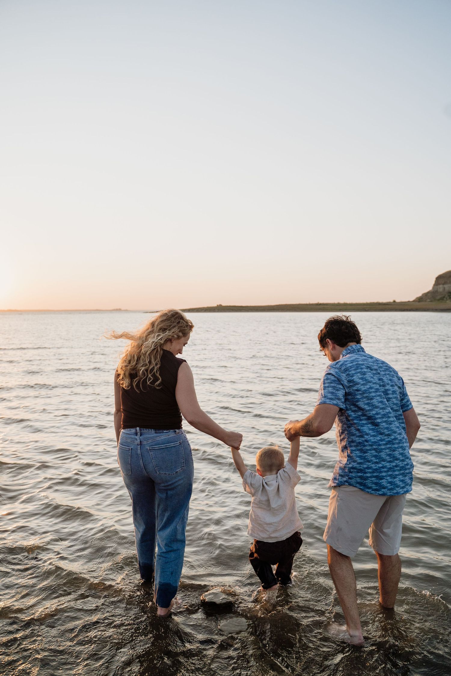 Family of three wading in shallow water at sunset, parents holding toddler's hands at a scenic lakeside.