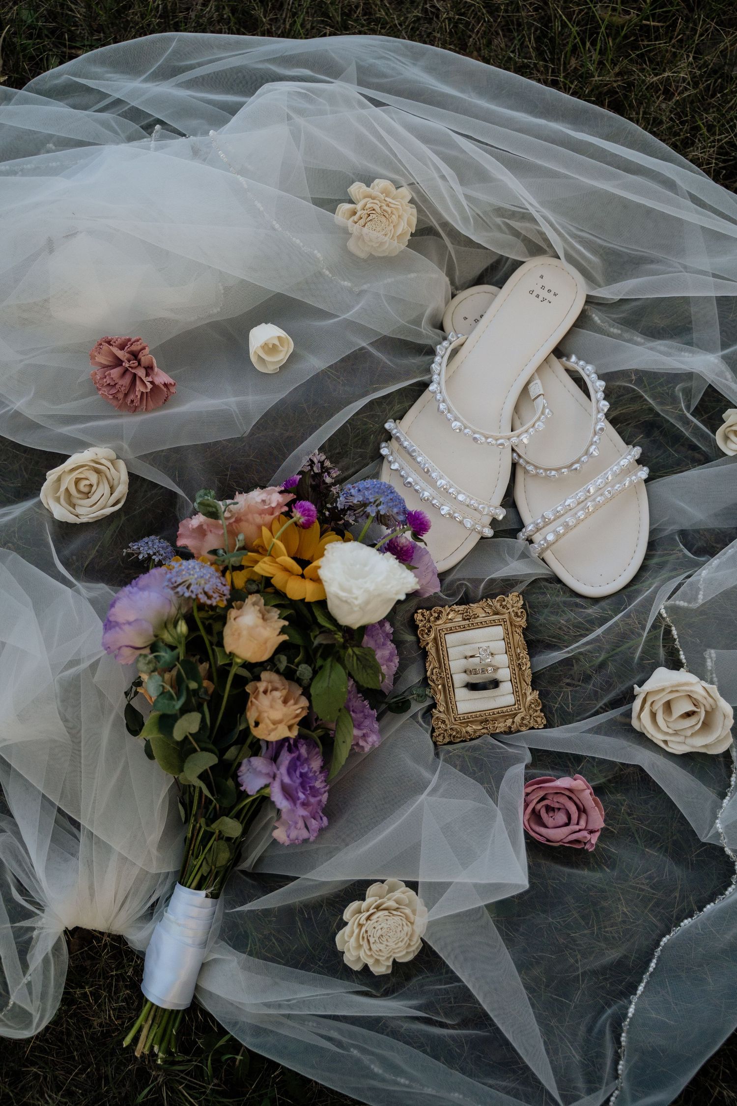 Flat lay of bridal details including pearl sandals, colorful bouquet, rings, and floral veil accents.