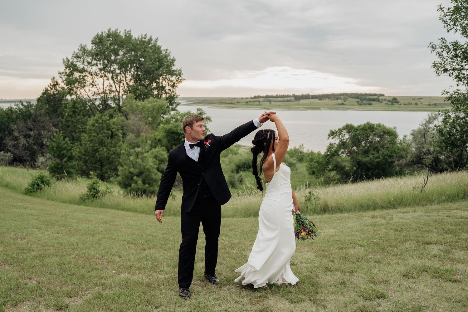 Groom twirling bride in white gown outdoors near a scenic lake on their wedding day.