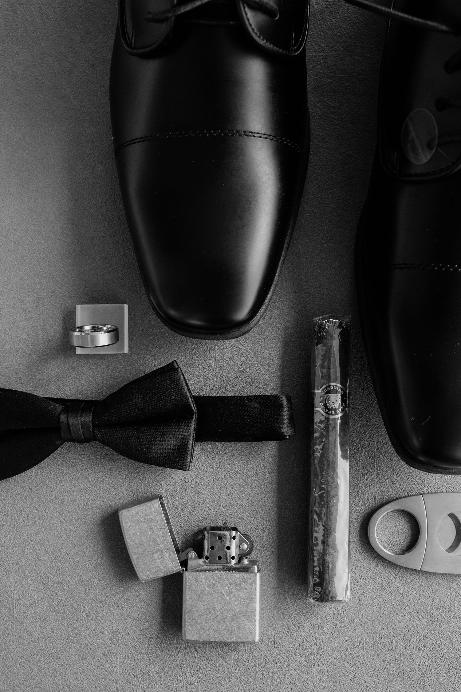Black and white flat lay of groom accessories including dress shoes, bow tie, cigar, lighter, and cufflinks.