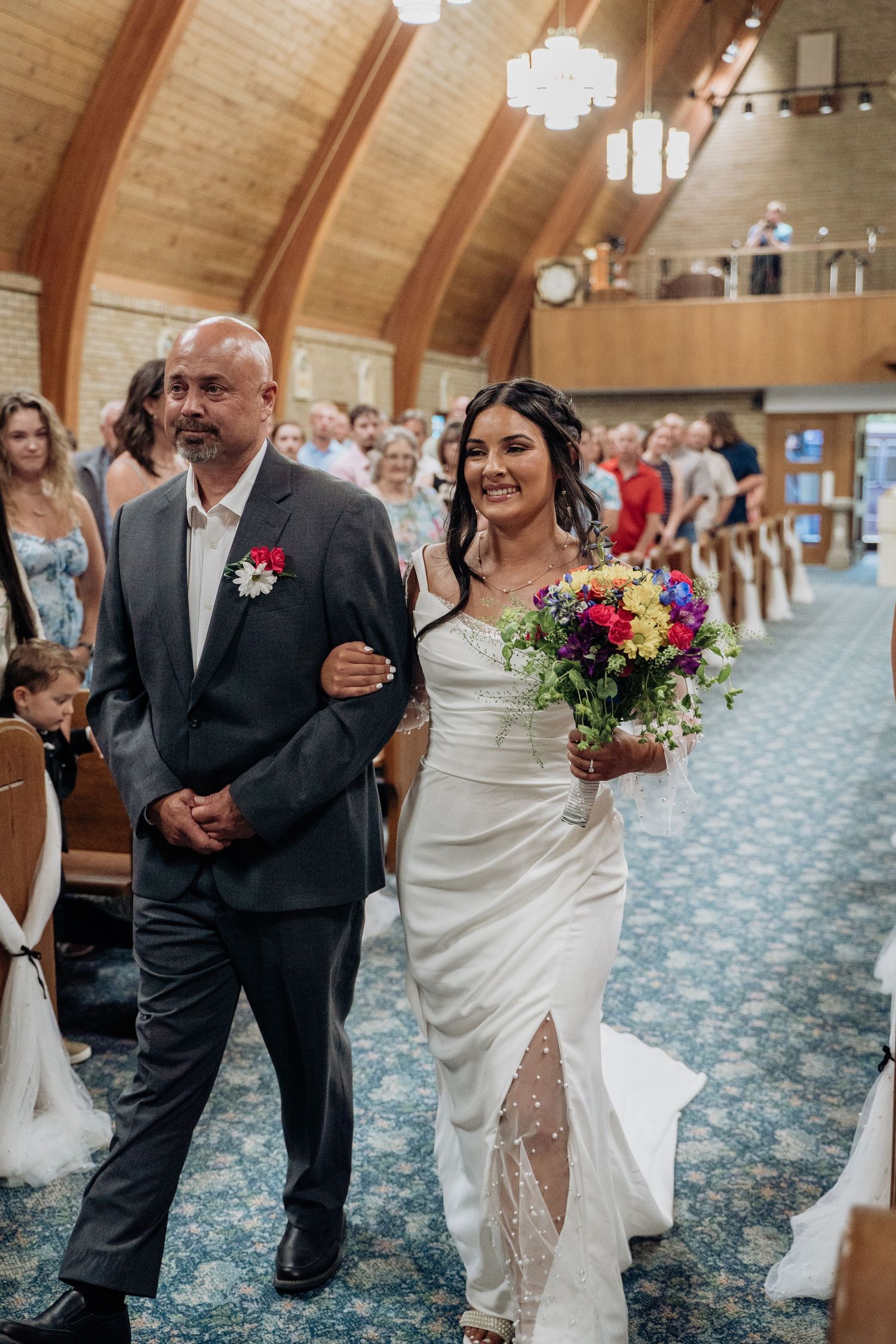 Bride walking down church aisle with father holding colorful bouquet during ceremony.